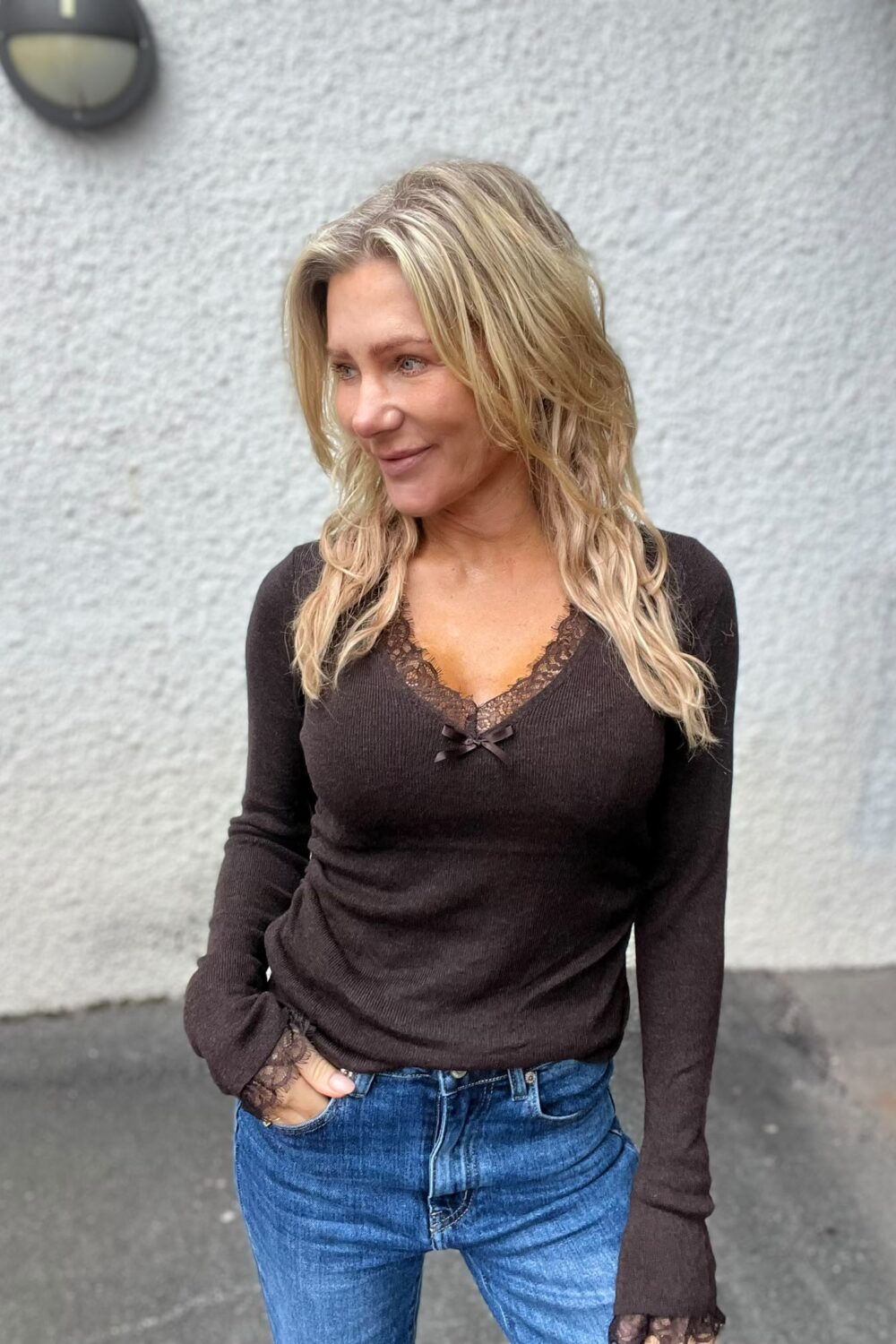 Blond kvinna står utomhus mot en ljusgrå putsad vägg, ler lätt och tittar åt vänster. Hon har ena handen i jeansfickan och bär en mörk, långärmad topp med spetsdetaljer vid ringning och ärmslut.
