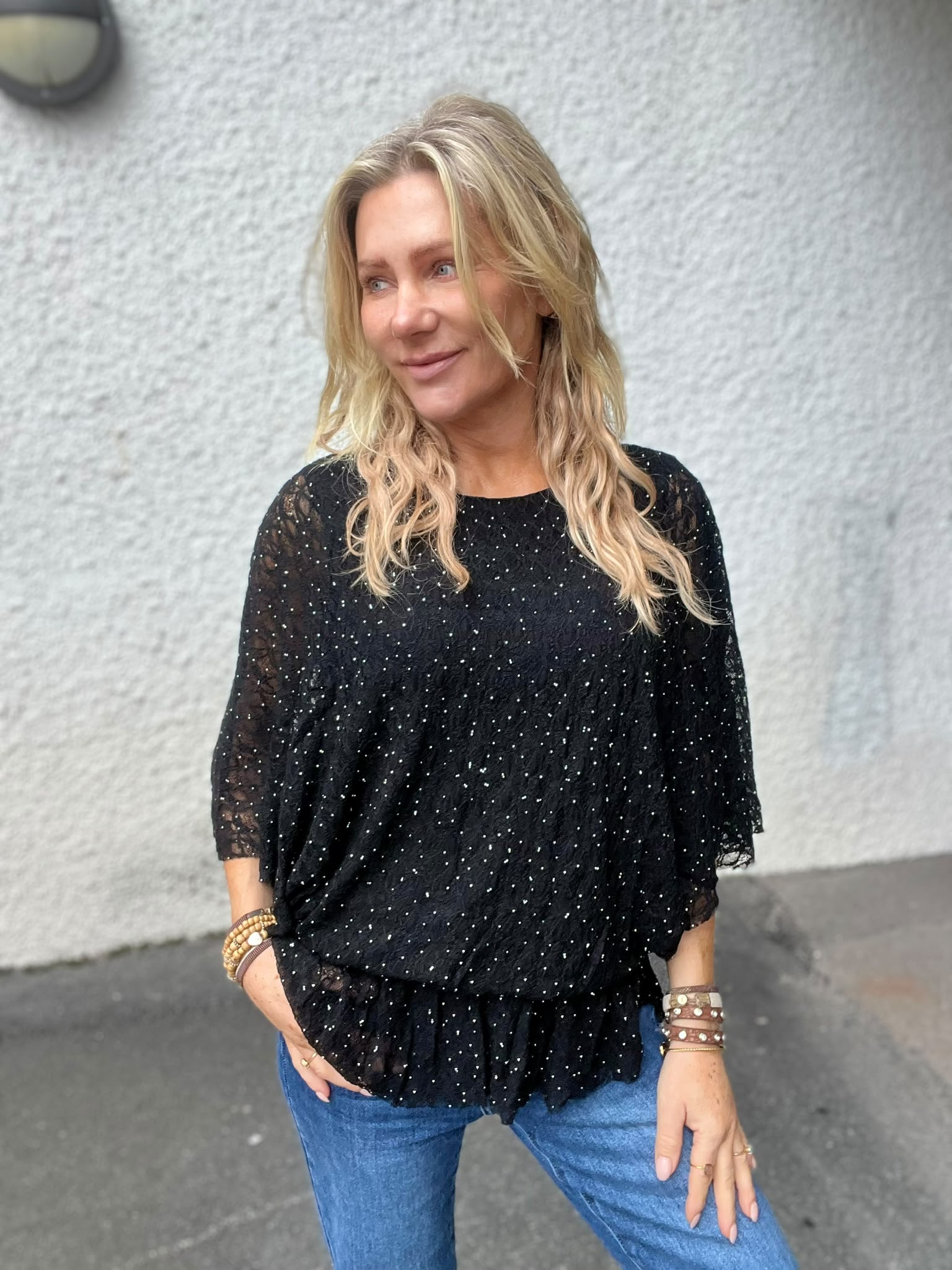 Blond kvinna står utomhus mot en ljus, grov vägg och tittar åt sidan med ett svagt leende. Hon bär svart glittrig spetsblus, blå jeans och flera armband, med ena handen i fickan.