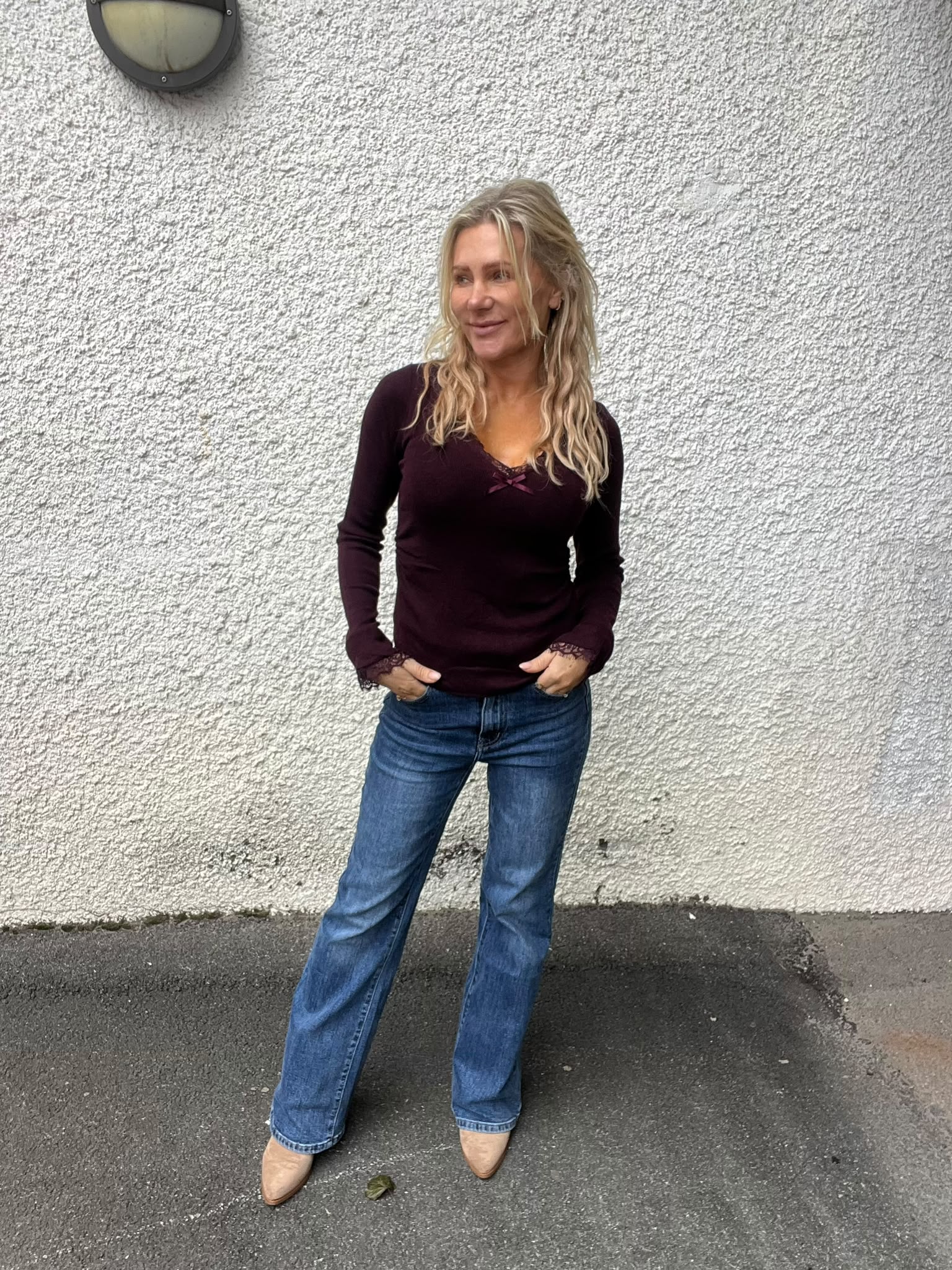Kvinna med långt blont hår står avslappnat med händerna i fickorna och tittar åt sidan. Hon bär mörklila långärmad topp och blå jeans med beige skor, framför en vit grovputsad vägg utomhus.