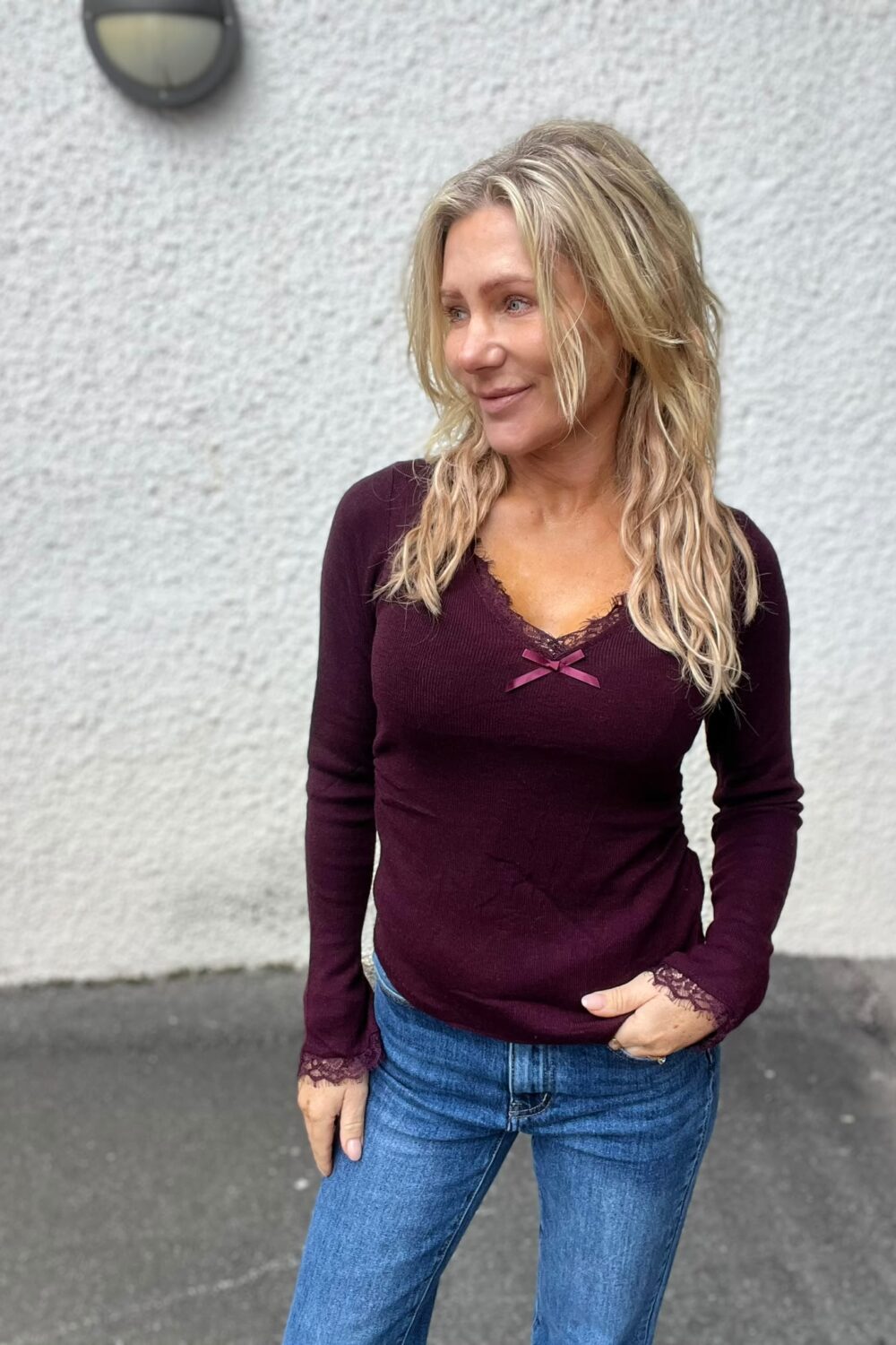 Blond kvinna står utomhus och tittar åt sidan med ett lätt leende. Hon har på sig vinröd långärmad topp med spetskant och liten rosett samt blå jeans, framför en ljus putsad vägg.