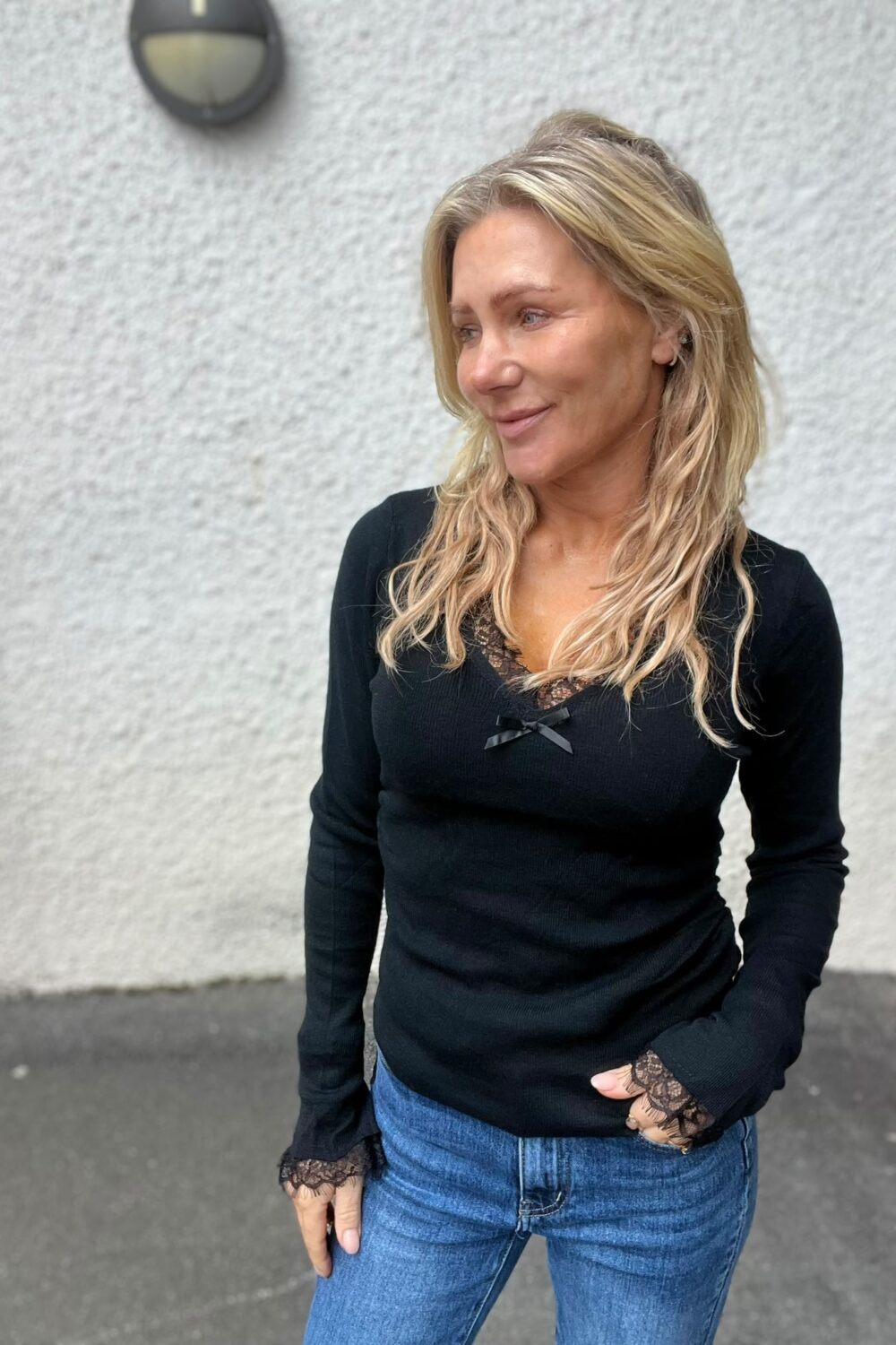 Blond kvinna står avslappnat med ena handen i jeansfickan och ler lätt medan hon tittar åt sidan. Hon bär svart långärmad topp med spetsdetaljer. Bakom henne syns en ljus, grov vägg och en rund vägglampa.