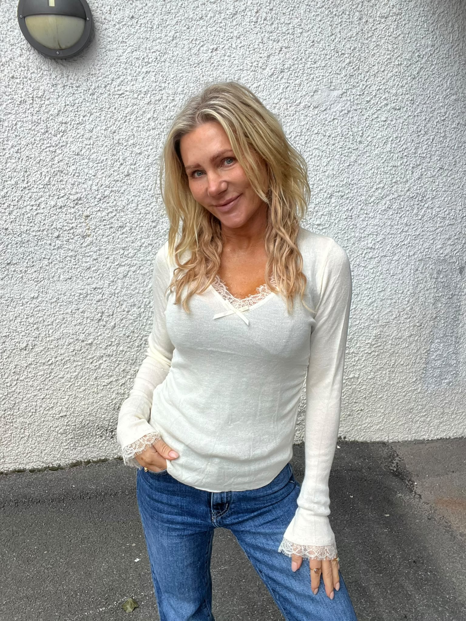 Blond kvinna står och ler mot kameran med en hand i jeansfickan och den andra på låret. Hon har vit långärmad topp med spetsdetaljer och står framför en ljus, grovputsad vägg med rund vägglampa.