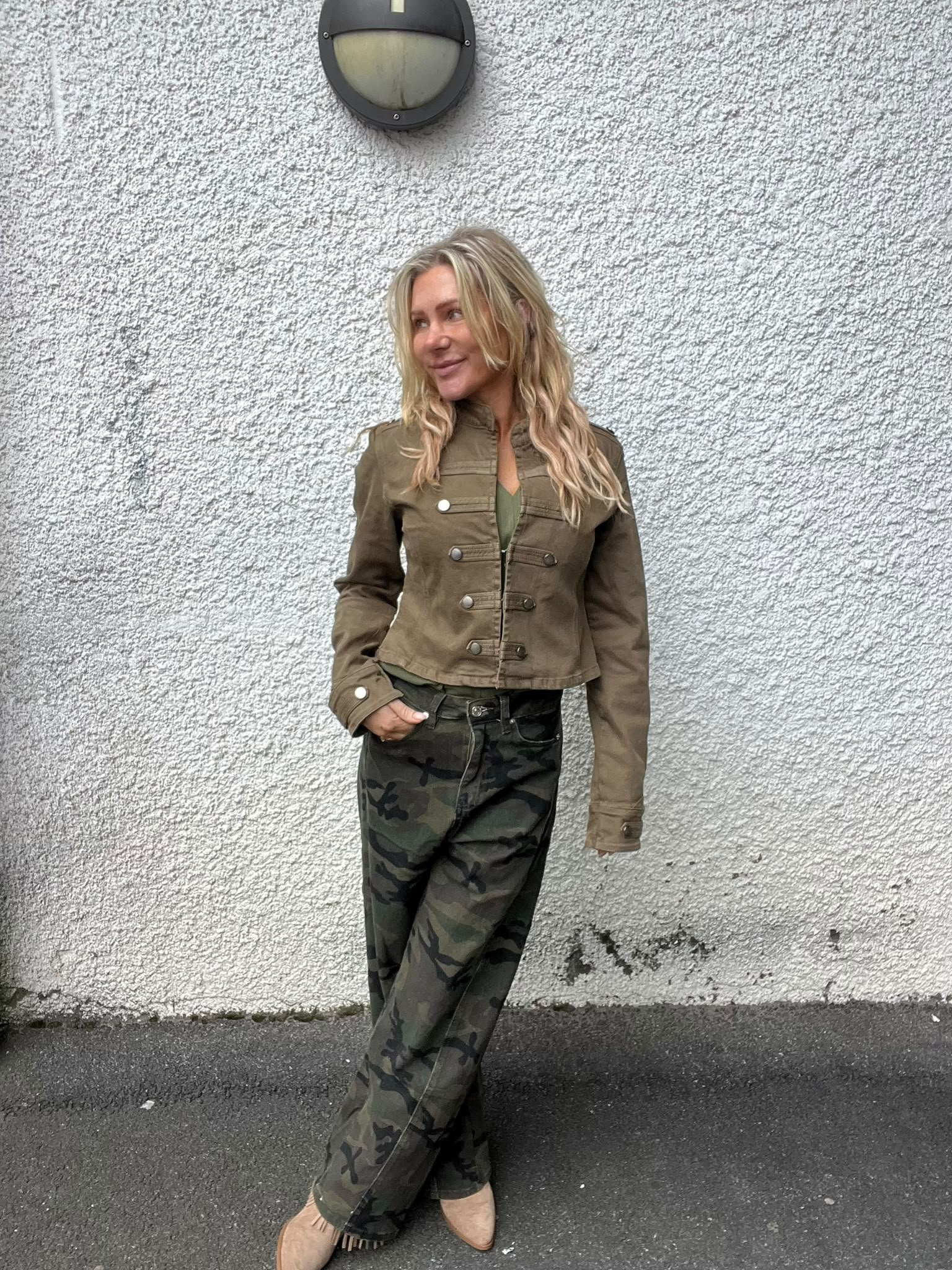 Blond kvinna står lutad mot en vit, grovputsad vägg och tittar åt sidan med ett leende. Hon har en olivgrön jacka, camouflagebyxor och beige skor; ovanför sitter en rund vägglampa.