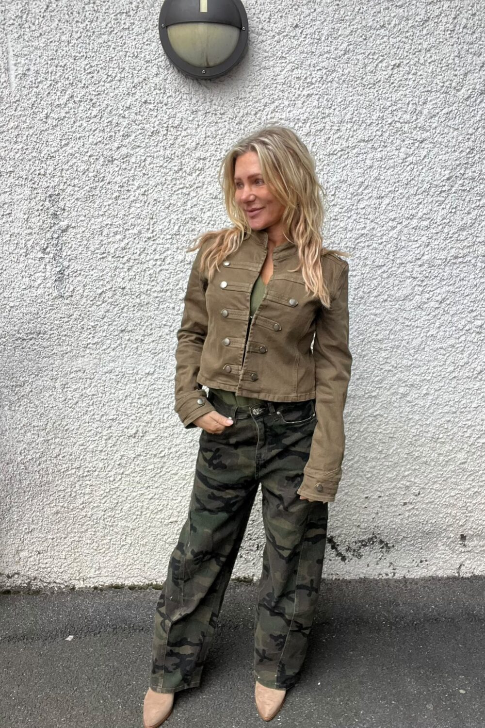 Blond kvinna står lutad mot en grov, vit yttervägg och tittar åt sidan med ett svagt leende. Hon bär kort brun jacka med knappar, kamouflagebyxor och beige boots; rund vägglampa ovanför.