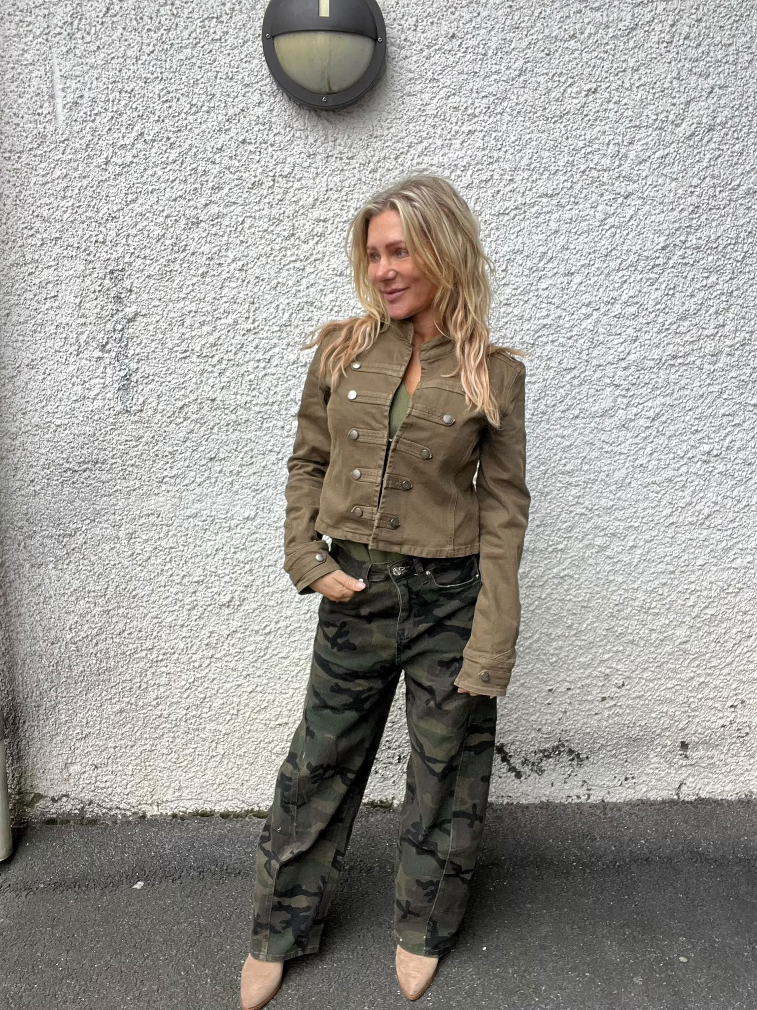 Blond kvinna står lutad mot en grov, vit yttervägg och tittar åt sidan med ett svagt leende. Hon bär kort brun jacka med knappar, kamouflagebyxor och beige boots; rund vägglampa ovanför.