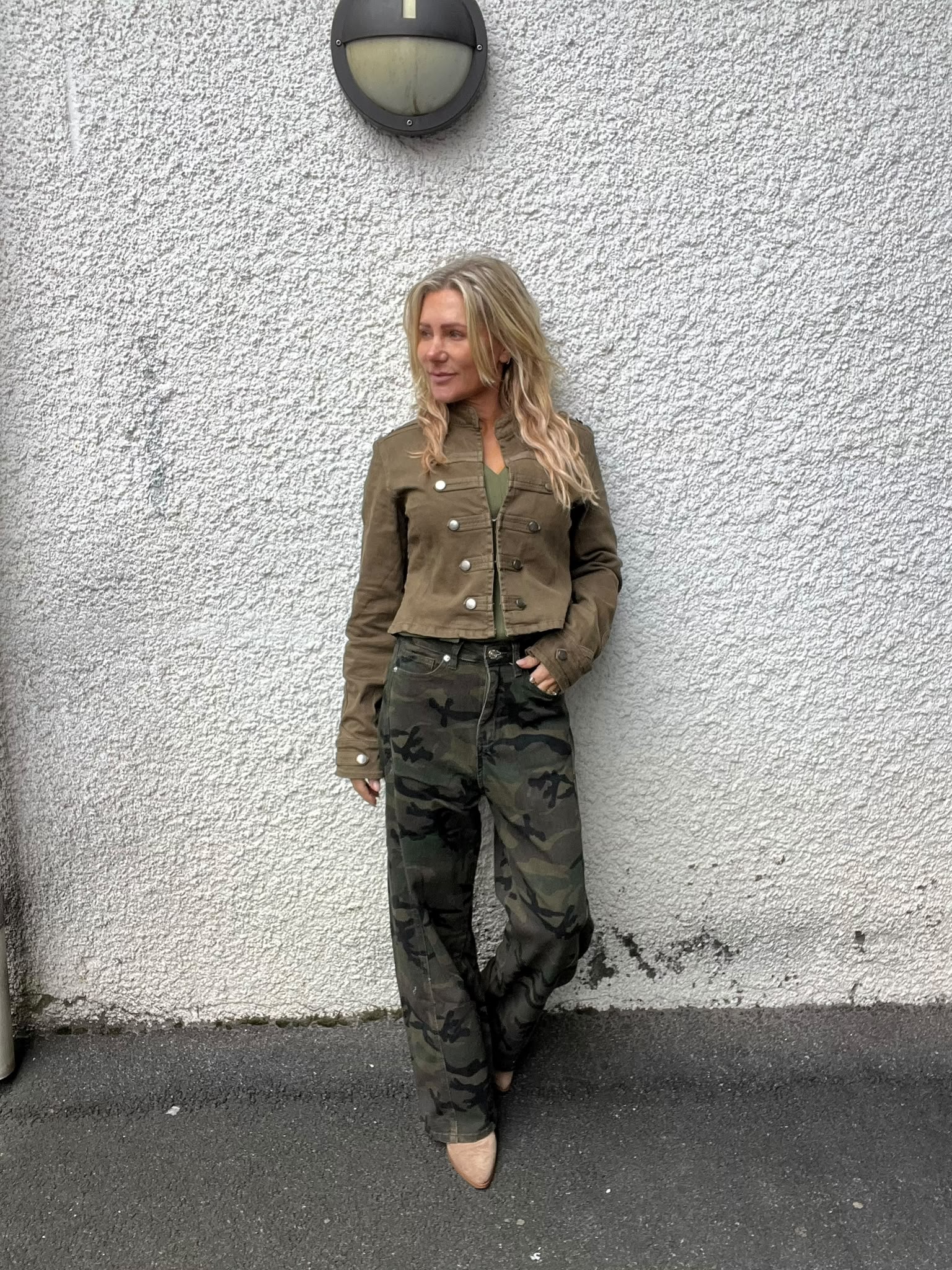 Blond kvinna står avslappnat lutad mot en vit, grovputsad vägg och tittar åt sidan. Hon bär kort brun jacka och kamouflagemönstrade byxor. Ovanför henne sitter en rund vägglampa, och hon står på mörk asfalt.