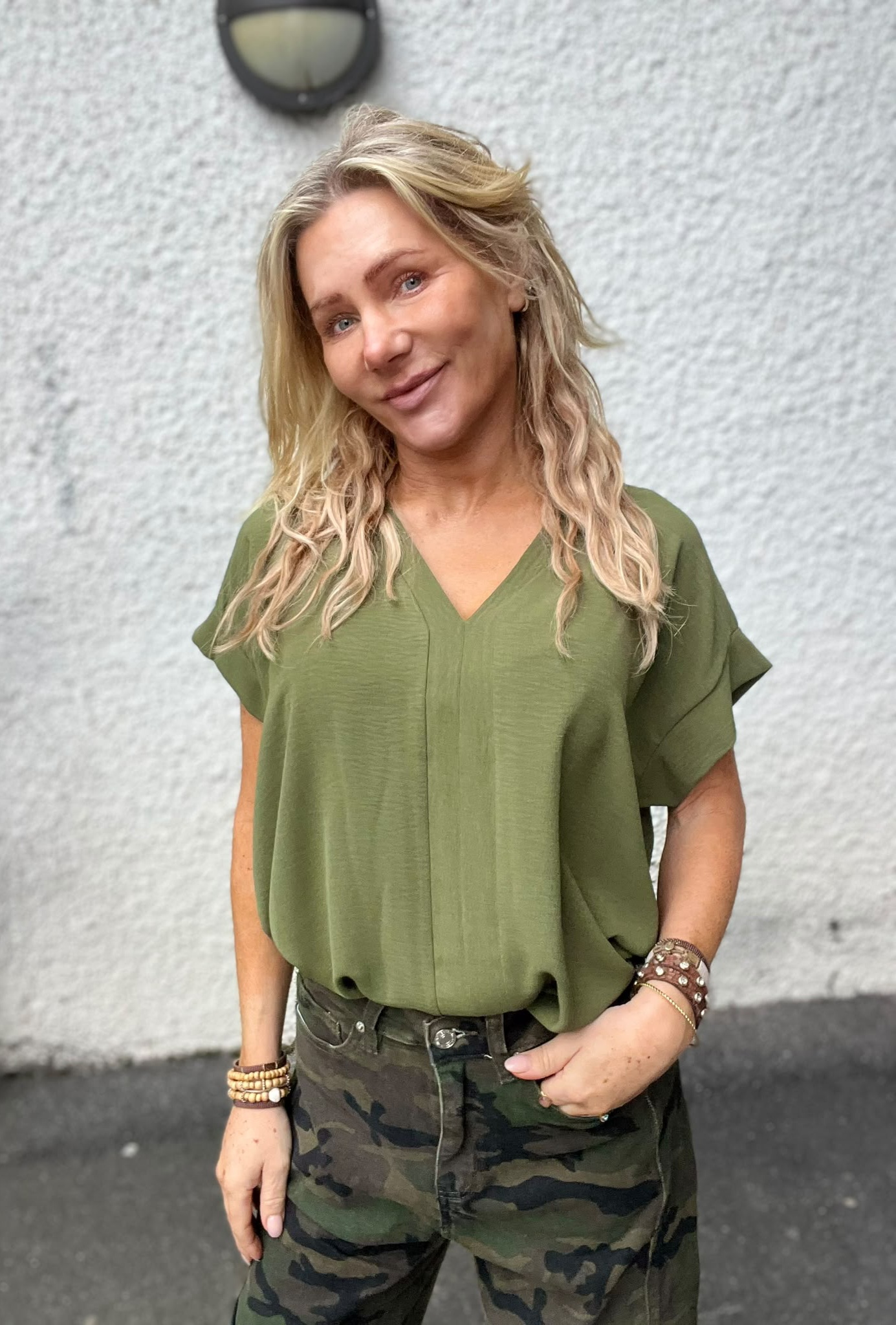 Leende kvinna med långt blont, vågigt hår står avslappnat med ena handen i fickan. Hon bär en olivgrön blus, kamouflagemönstrade byxor och flera armband, framför en ljus, grovputsad vägg med vägglampa.