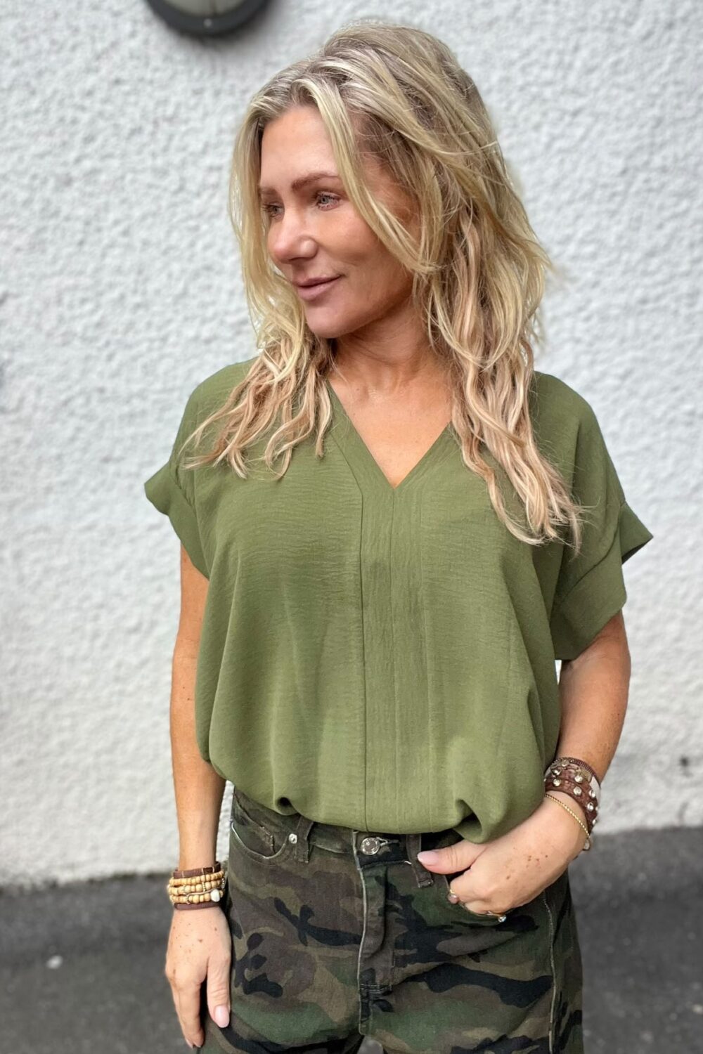 Blond kvinna står utomhus och tittar åt vänster med ett svagt leende. Hon bär en olivgrön V-ringad blus, kamouflagebyxor och flera armband. Bakom henne syns en ljus, grov putsad vägg.
