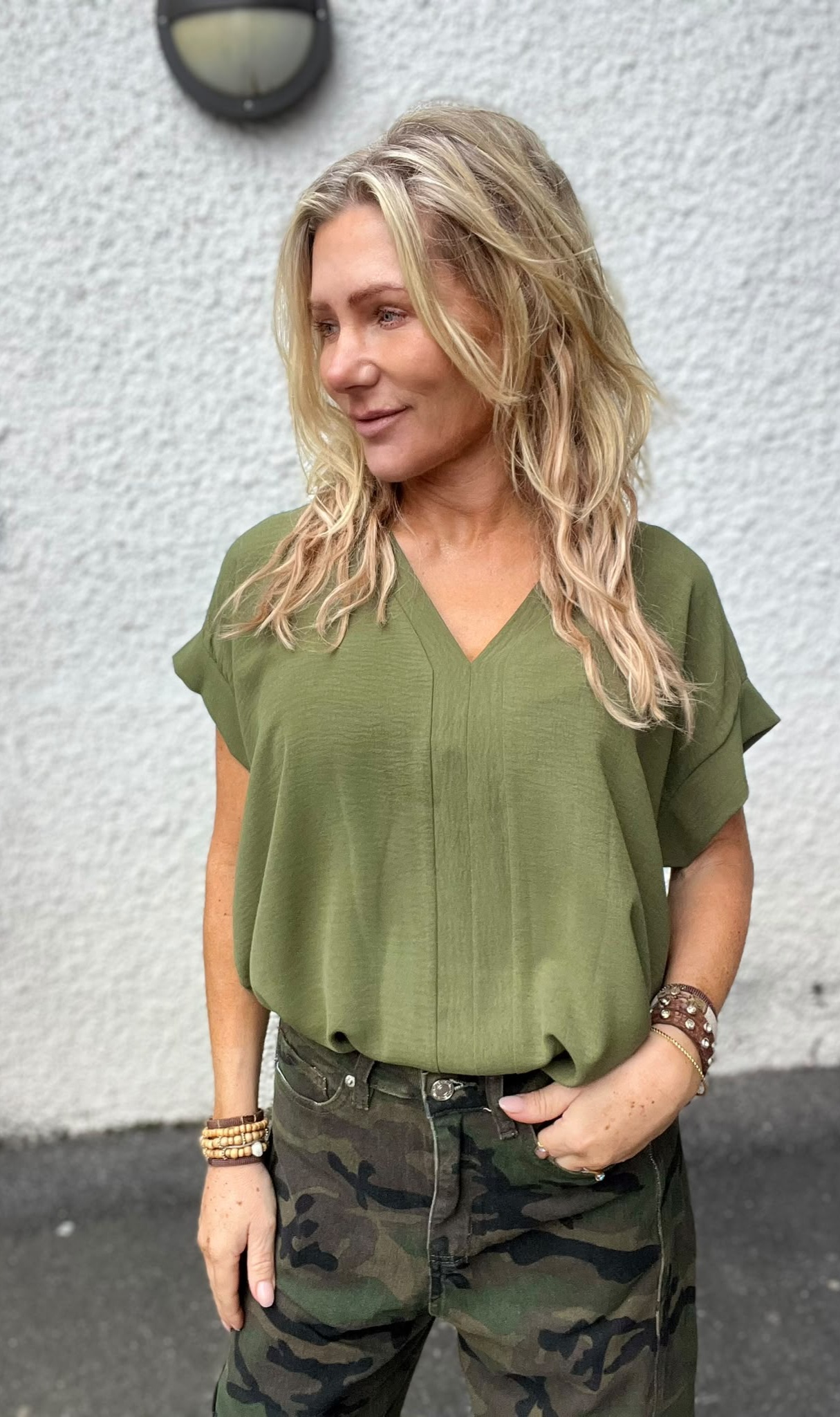 Blond kvinna står utomhus och tittar åt vänster med ett svagt leende. Hon bär en olivgrön V-ringad blus, kamouflagebyxor och flera armband. Bakom henne syns en ljus, grov putsad vägg.