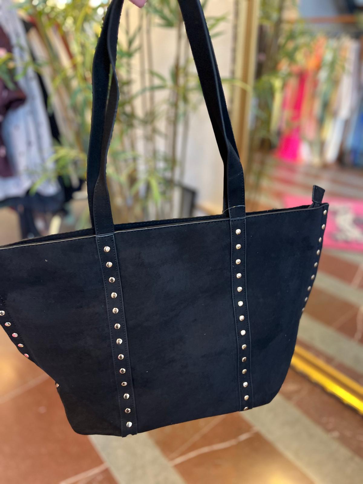 Svart totebag i mockaliknande material hänger i två axelremmar, dekorerad med silverfärgade nitar längs vertikala band och sidor. Fotograferad nära en butiksmiljö med kläder och växter suddigt i bakgrunden.