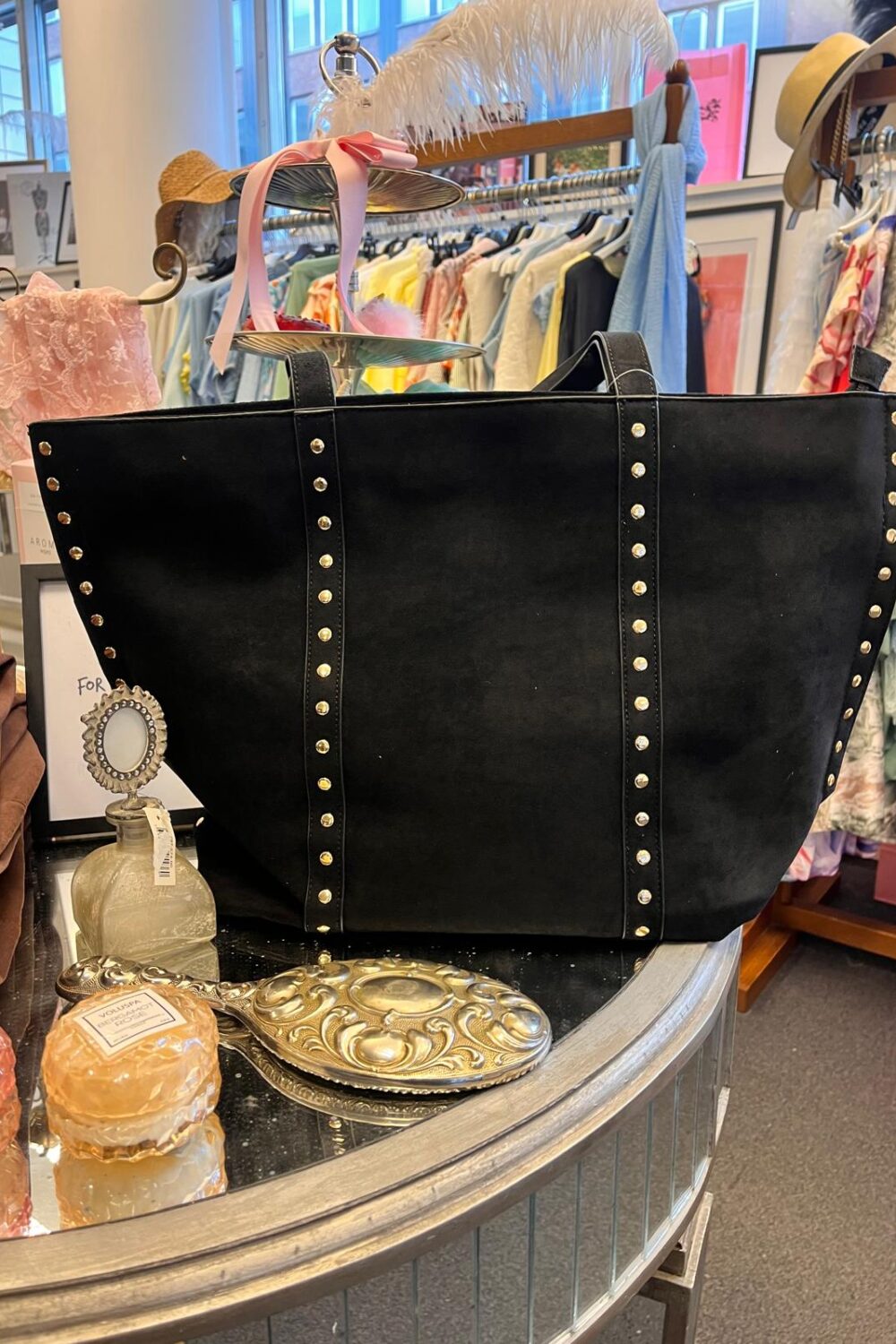 Stor svart toteväska med nitar står uppställd på ett runt bord i en klädbutik, omgiven av parfymflaska, tvål och en dekorativ borste. I bakgrunden syns klädställning med färgglada plagg och hattar.
