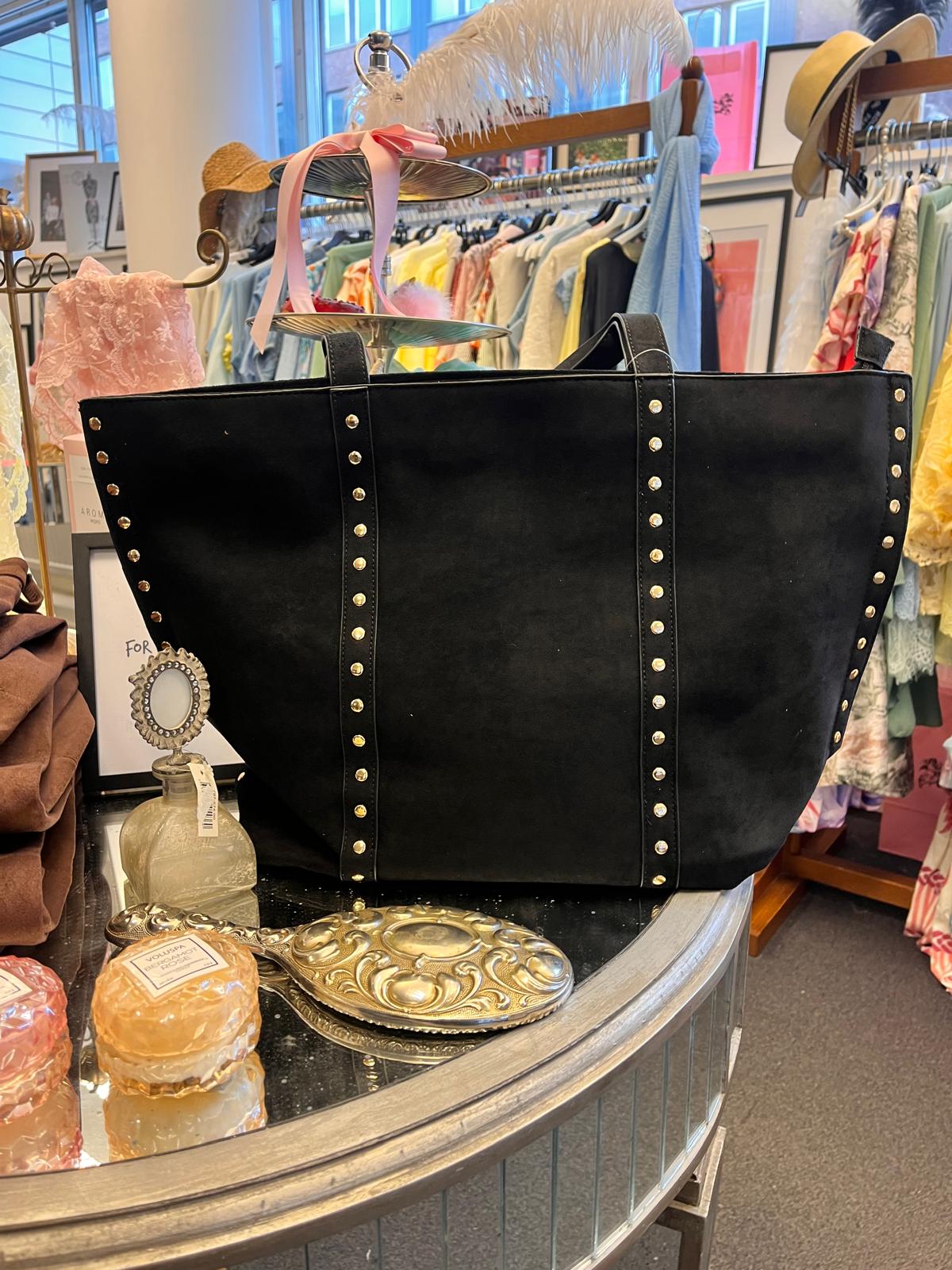 Stor svart toteväska med nitar står uppställd på ett runt bord i en klädbutik, omgiven av parfymflaska, tvål och en dekorativ borste. I bakgrunden syns klädställning med färgglada plagg och hattar.