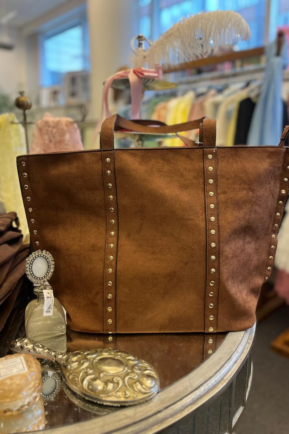 Stor brun mockatote med nitar och dubbla handtag står uppställd på ett runt bord i en butik. I förgrunden syns små prydnader och en flaska; i bakgrunden hänger kläder och en fjäderdekoration.