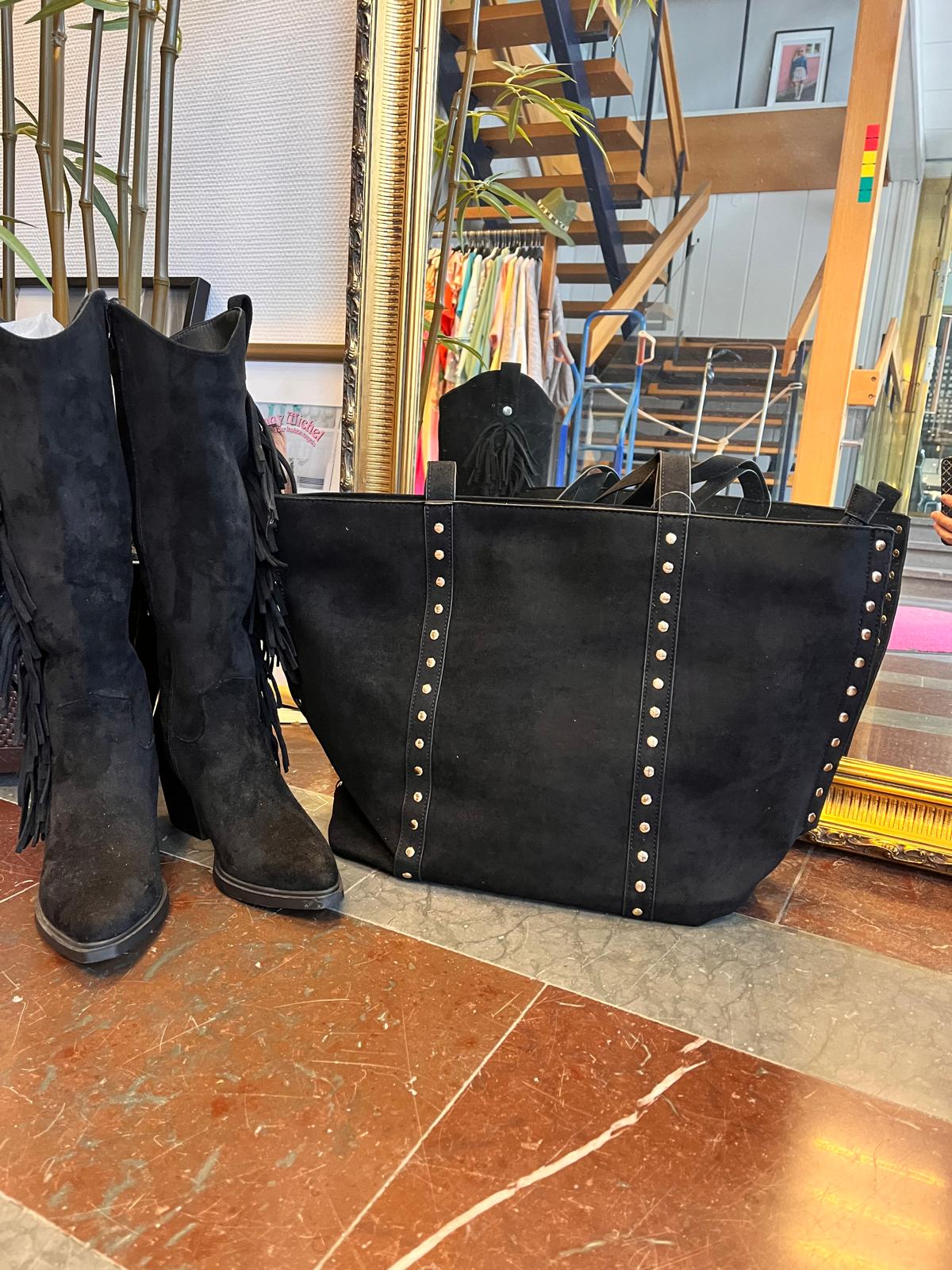 Svarta mockastövlar med fransar står lutade mot en stor svart tote-väska med nitar och dubbla handtag. Produkterna är uppställda på ett marmorgolv i en butik, med spegel, trappa och klädställning i bakgrunden.