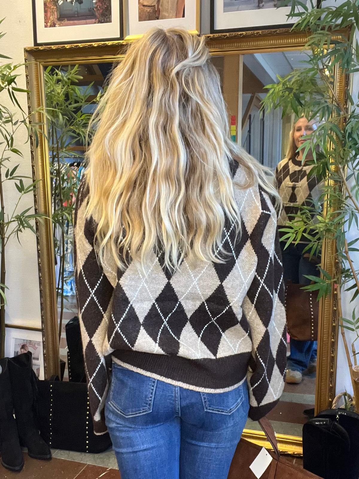 Kvinna med långt blont, vågigt hår står med ryggen mot kameran och tittar i en stor guldramsspegel; hennes ansikte syns i spegeln. Hon bär en brun-beige argyletröja och jeans, omgiven av gröna växter inomhus.