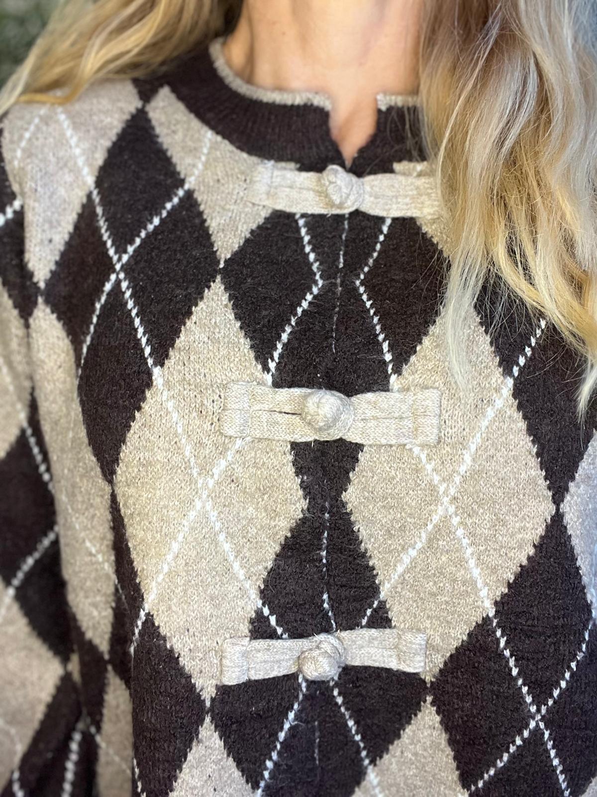 Närbild av en person som bär en stickad kofta med argylemönster i svart, beige och vitt. Koftan är knäppt med tre ljusa toggleknäppningar framtill, och blont hår faller över ena axeln.