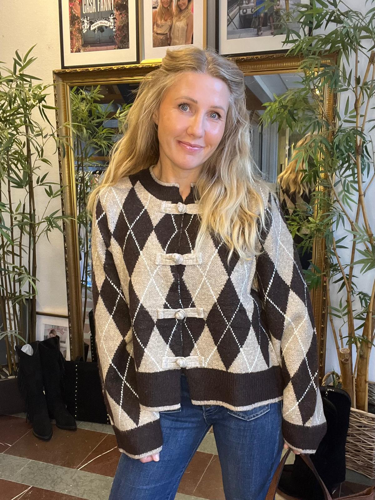 Kvinna med långt blont hår står och ler mot kameran, iklädd en brun- och beigefärgad argyle-stickad tröja med dekorativa knappar och jeans, inomhus framför en stor spegel, gröna växter och inramade foton på väggen.