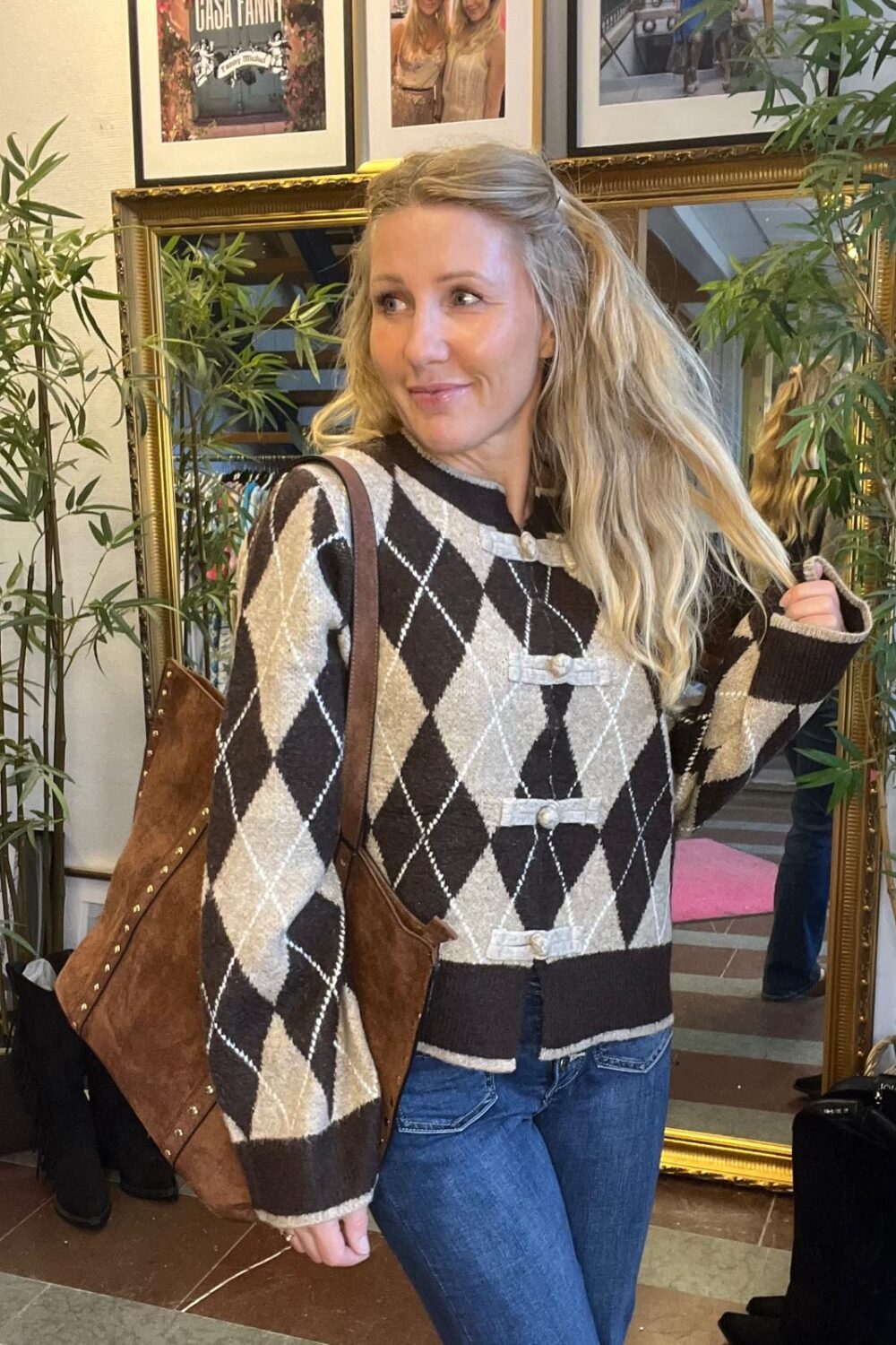 Blond kvinna står inomhus och tittar åt sidan med ett leende, iklädd brun‑beige argylemönstrad kofta och jeans, med stor brun axelväska. Bakom henne syns en guldramsspegel, bambuväxter och inramade foton.