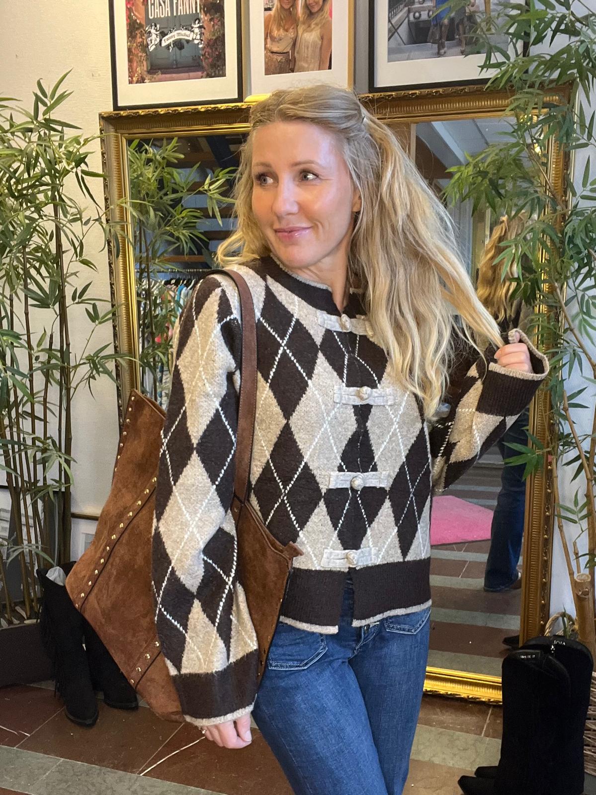 Blond kvinna står inomhus och tittar åt sidan med ett leende, iklädd brun‑beige argylemönstrad kofta och jeans, med stor brun axelväska. Bakom henne syns en guldramsspegel, bambuväxter och inramade foton.