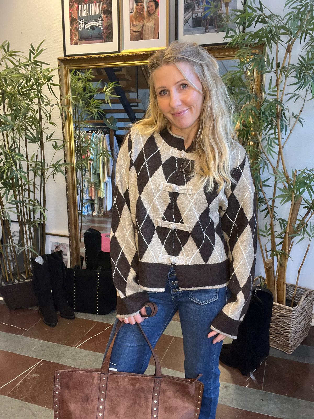 Leende kvinna står inomhus och håller en stor brun väska. Hon bär en beige och brun argylemönstrad kofta och jeans. Bakom henne syns spegel, klädställning, stövlar och väggbilder med texten ”CASA FANNY”.