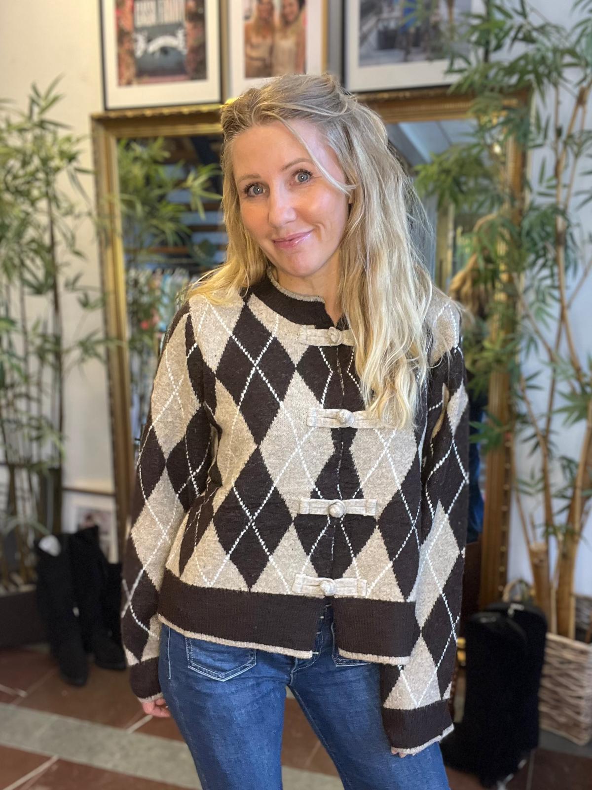 Blond kvinna ler mot kameran och står avslappnat inomhus, klädd i brun-beige argylemönstrad kofta med knäppningar och blå jeans. Bakgrunden visar en stor spegel, krukväxter och inramade foton på väggen.