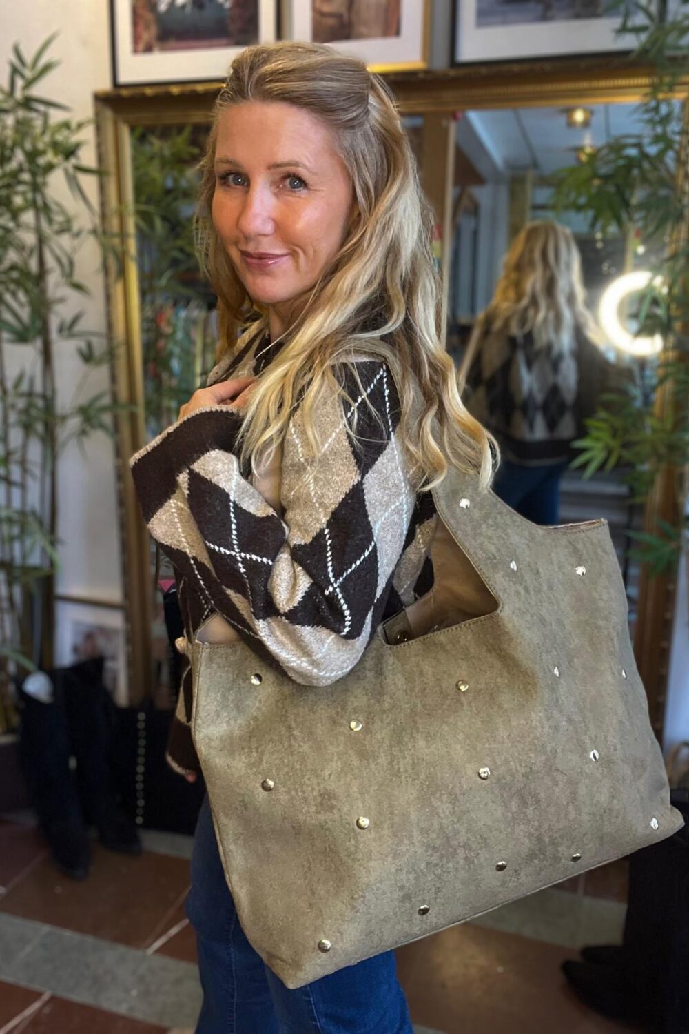 Blond kvinna poserar med en stor grå mockaväska med nitar över axeln och tittar mot kameran. Hon bär en brun- och beige-rutig stickad tröja, i en butiksmiljö med spegel och gröna växter.