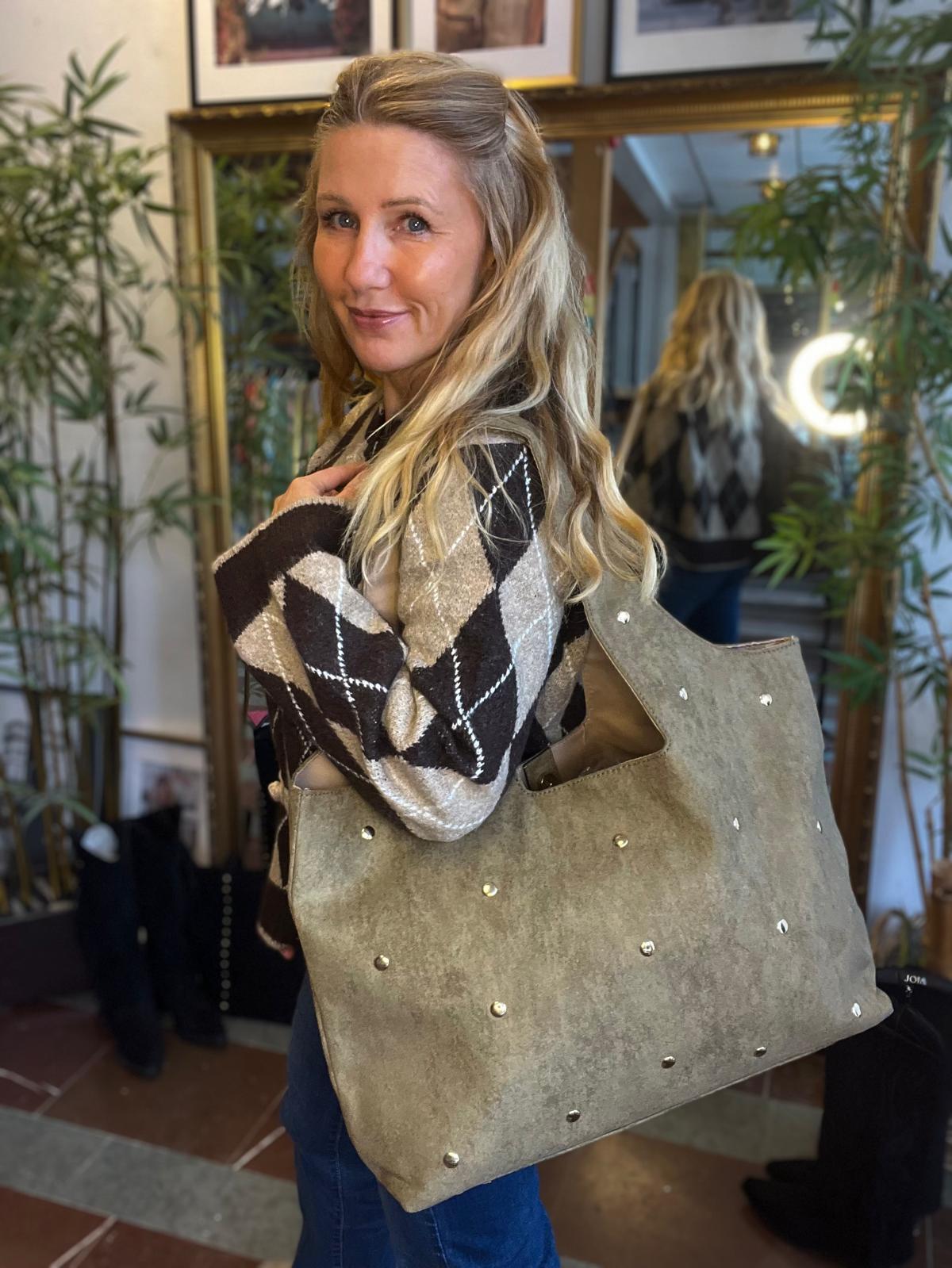 Blond kvinna poserar med en stor grå mockaväska med nitar över axeln och tittar mot kameran. Hon bär en brun- och beige-rutig stickad tröja, i en butiksmiljö med spegel och gröna växter.