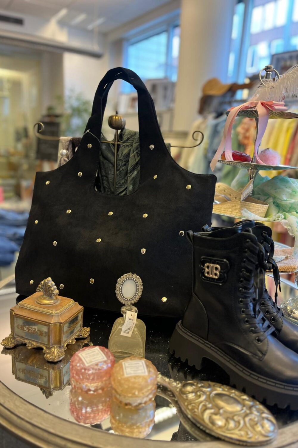 Svart handväska med nitar står i fokus på ett bord i en butik, bredvid svarta snörkängor. Runtom ligger en liten parfymflaska med prislapp, dekorativa askar och ett smyckesfat; kläder syns suddigt i bakgrunden.