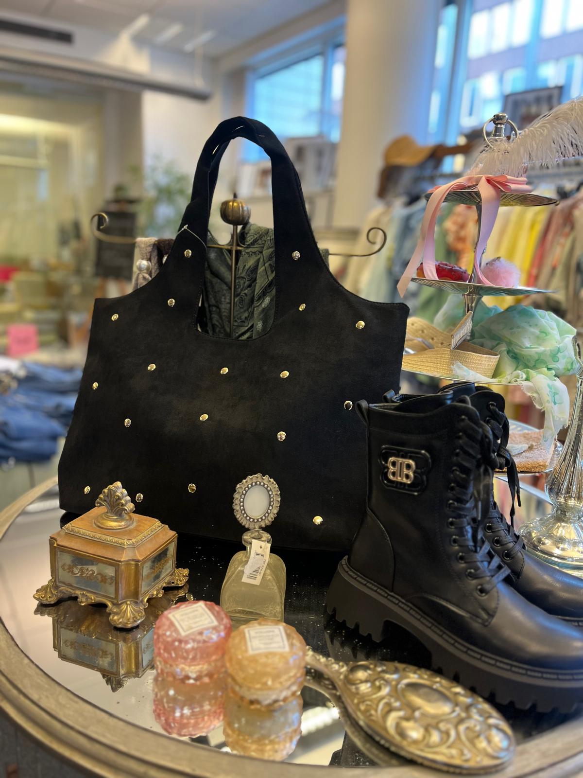 Svart handväska med nitar står i fokus på ett bord i en butik, bredvid svarta snörkängor. Runtom ligger en liten parfymflaska med prislapp, dekorativa askar och ett smyckesfat; kläder syns suddigt i bakgrunden.