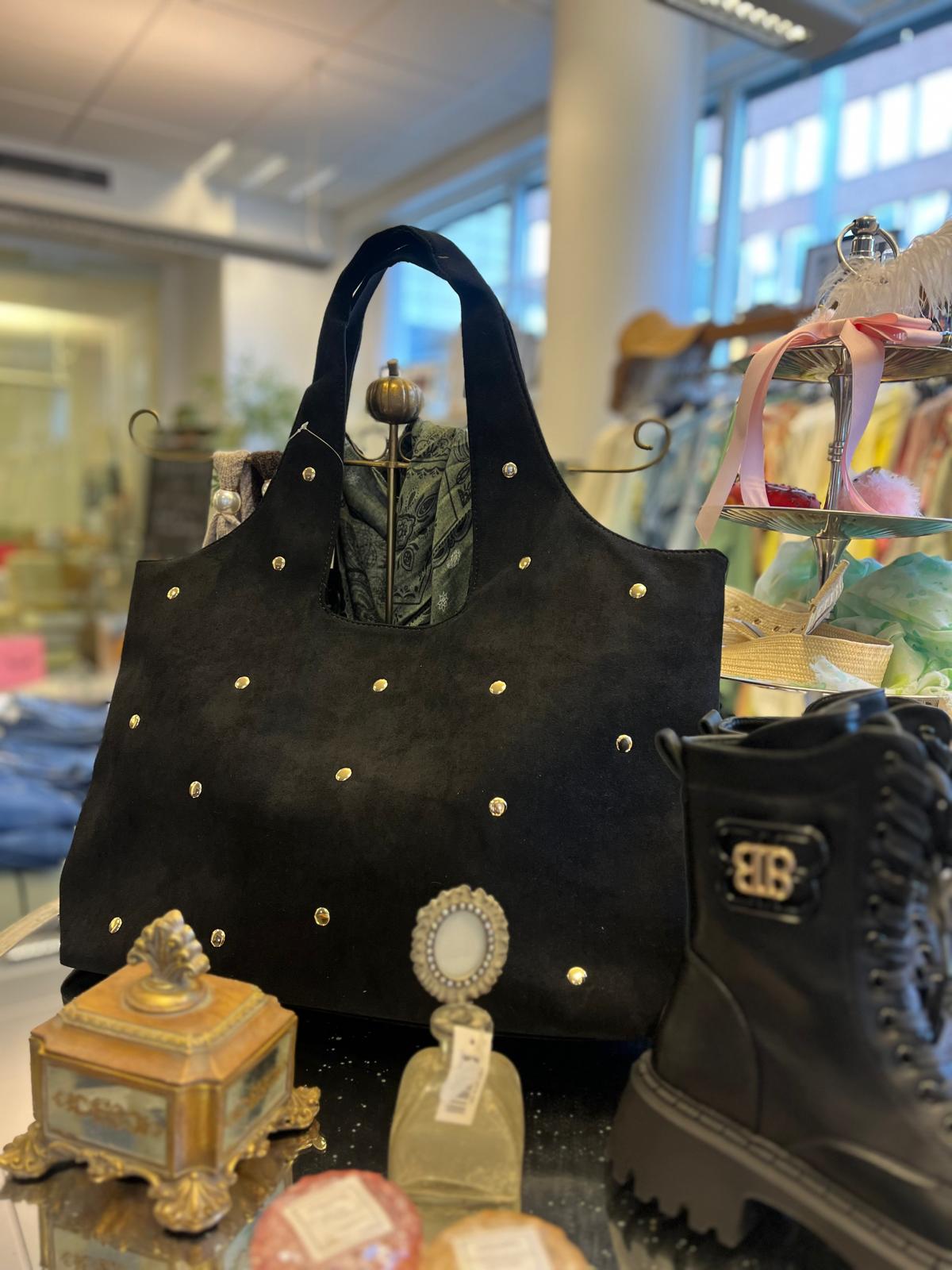 Stor svart handväska i mockaliknande material med guldfärgade nitar står på ett bord i en butik, med en sjal instoppad. Intill syns svart känga med “BB”-spänne och små prydnadsföremål.