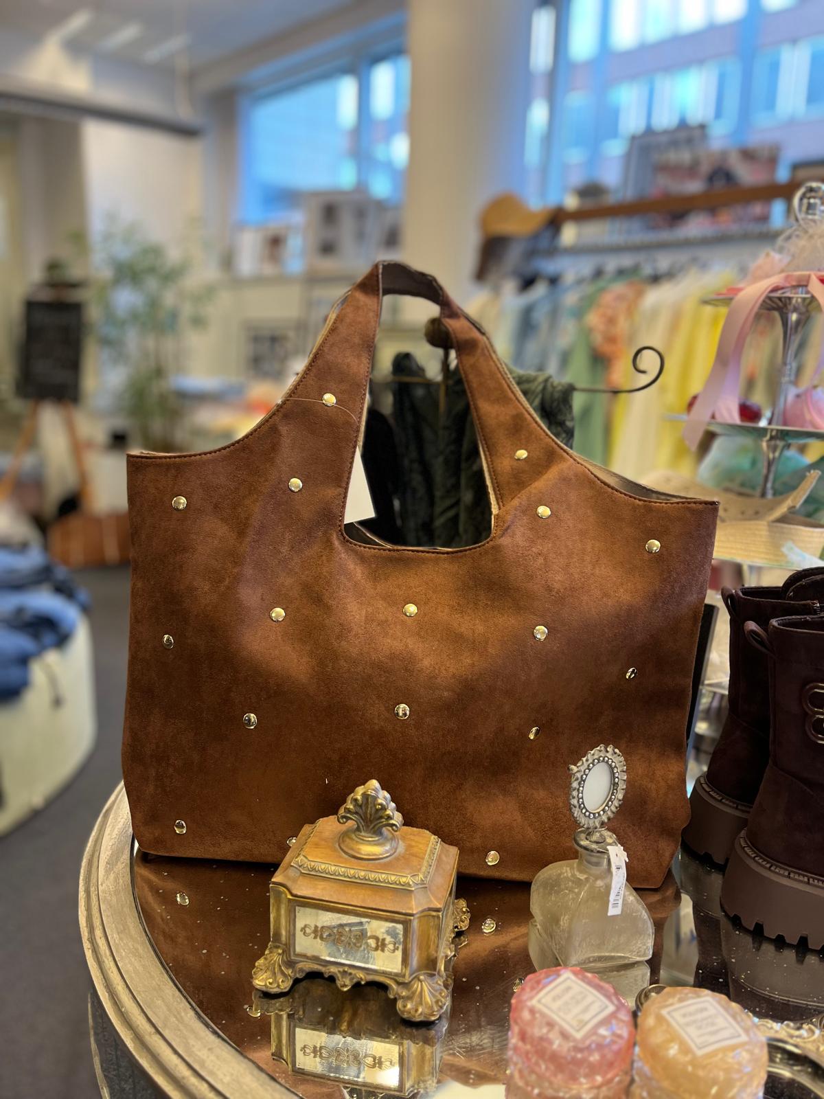 Brun mockaväska med nitar står uppställd på ett runt bord i en butik. Framför syns små dekorativa föremål och tvålar, och till höger står mörka kängor; i bakgrunden hänger kläder på ställningar.