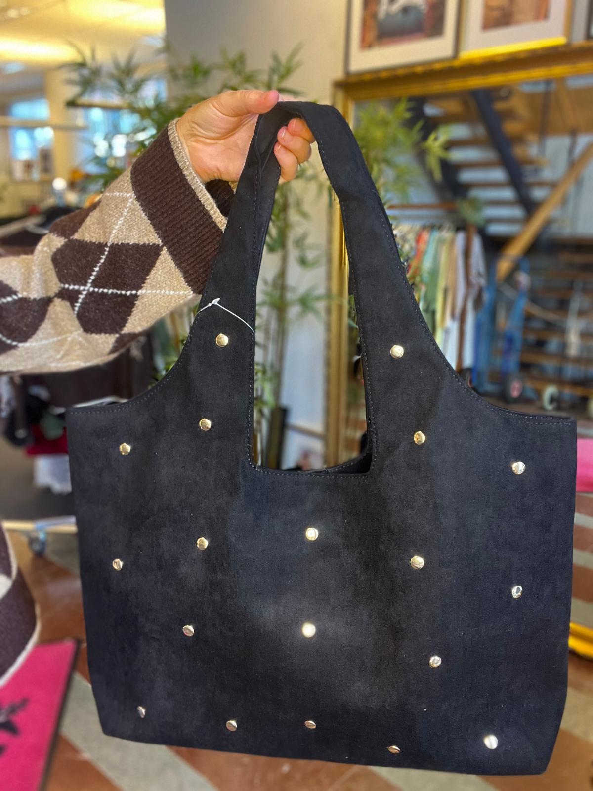 Hand håller upp en svart, mjuk tyg- eller mockaväska med två handtag och små metallnitar som prickar. Väskan visas i närbild inomhus, med suddig bakgrund av butiksmiljö och trappa.