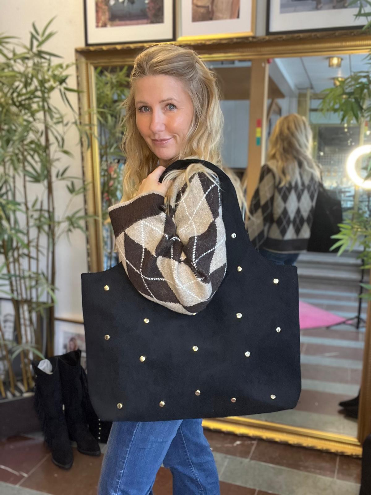 Blond kvinna poserar inomhus och tittar mot kameran medan hon bär en stor svart totebag med guldfärgade nitar på axeln. Hon står framför en stor guldramsspegel, med gröna växter och inramade bilder i bakgrunden.