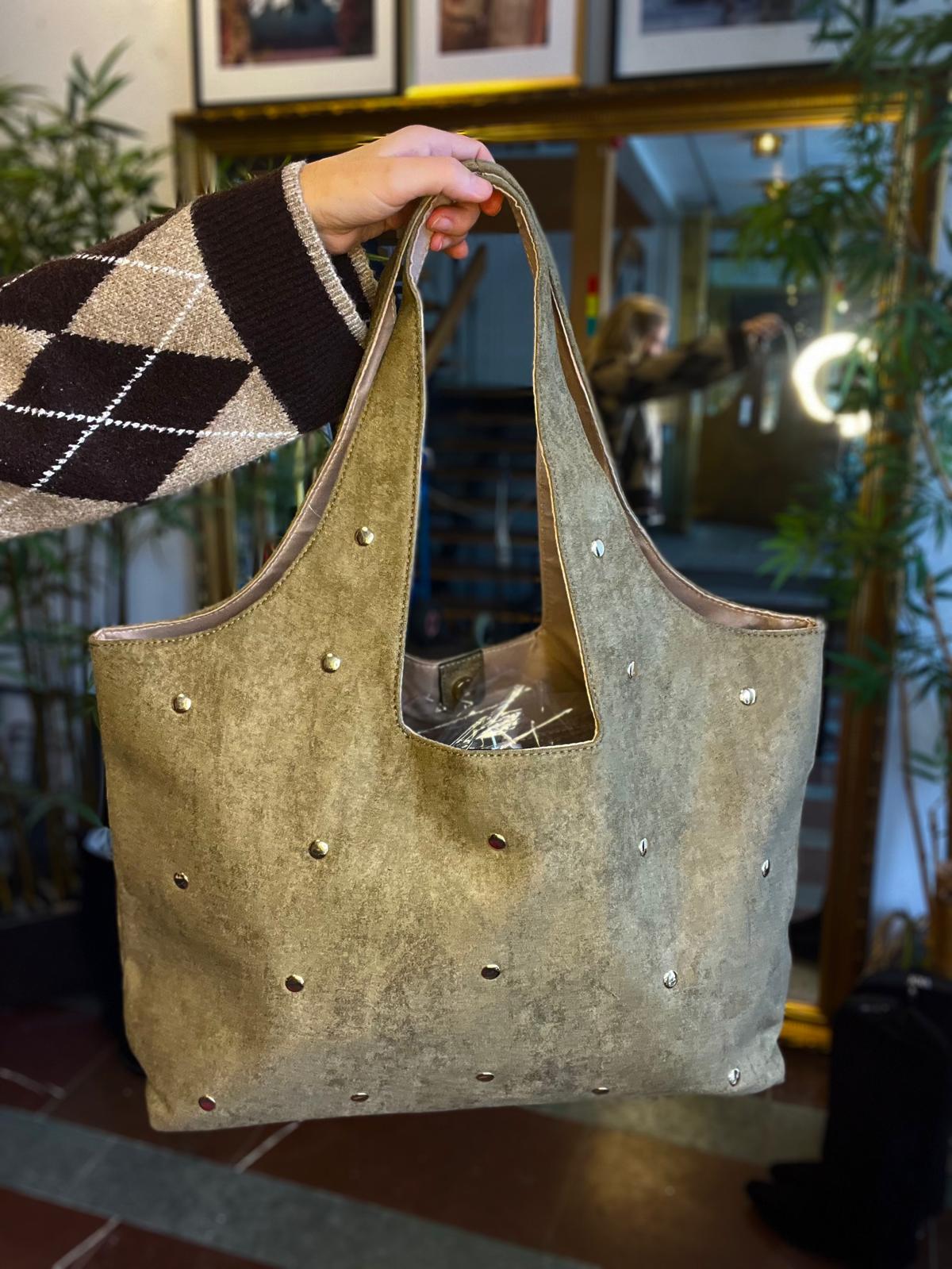 Hand i rutmönstrad tröja håller upp en stor grå-beige mockaväska med breda handtag och små metallnitar. Väskan visas inomhus; i bakgrunden syns spegel, växter och en ringlampa.