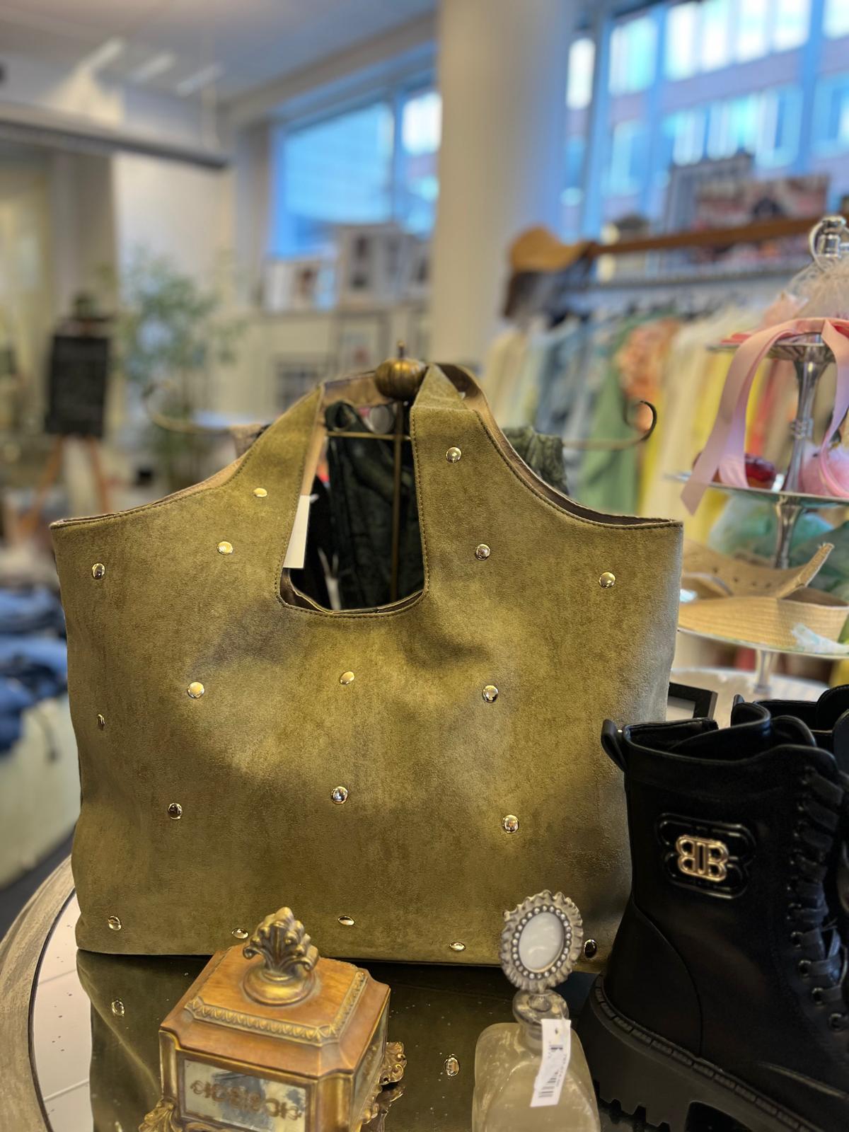 Olivgrön mockaväska med nitar står uppställd på ett bord i en butik. Bredvid syns svarta kängor och små dekorföremål, medan klädställningar och stora fönster är suddiga i bakgrunden.
