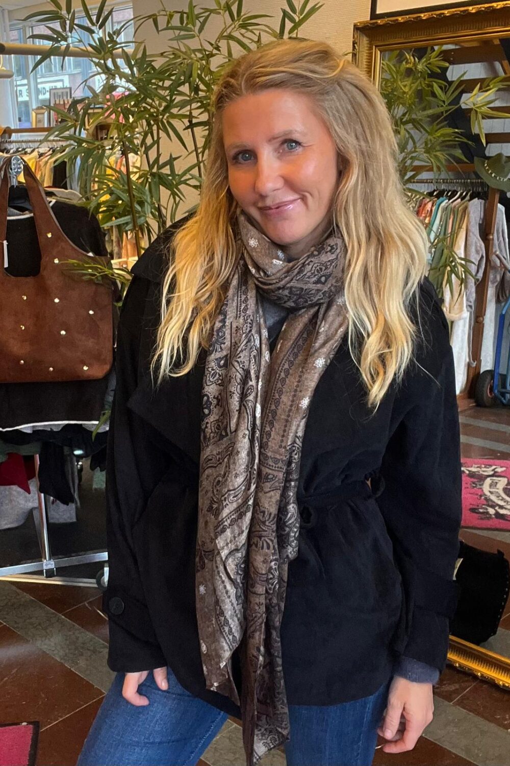 Leende kvinna med långt blont hår står och poserar med händerna i fickorna, iklädd svart kappa och mönstrad sjal. Hon befinner sig i en klädbutik med klädställningar, växter och en brun läderväska i bakgrunden.