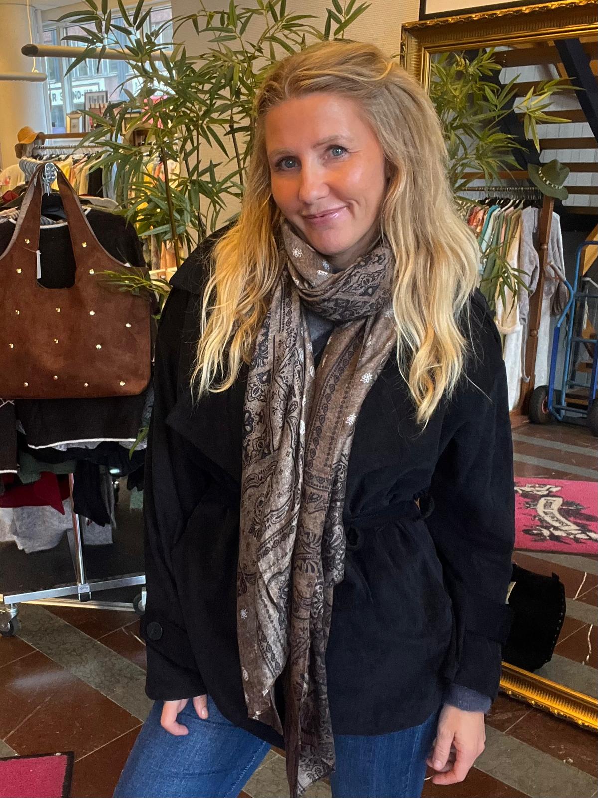 Leende kvinna med långt blont hår står och poserar med händerna i fickorna, iklädd svart kappa och mönstrad sjal. Hon befinner sig i en klädbutik med klädställningar, växter och en brun läderväska i bakgrunden.
