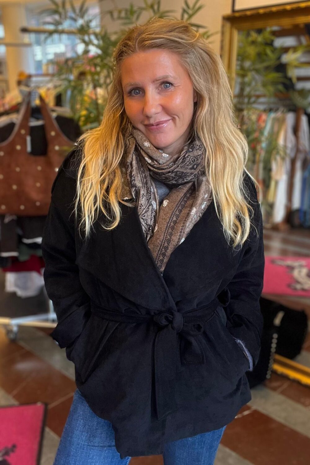 Blond kvinna står och ler mot kameran med händerna i fickorna, klädd i svart knuten jacka, mönstrad halsduk och jeans. Hon är inomhus i en butik med klädställningar och växter i bakgrunden.