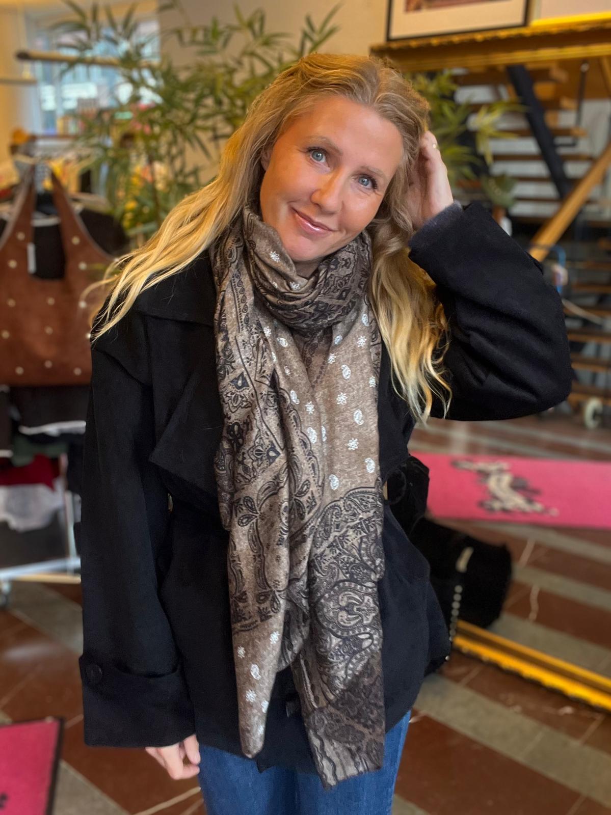 Leende kvinna med långt blont hår står inomhus och lutar huvudet mot ena handen. Hon bär svart jacka och en stor mönstrad sjal. I bakgrunden syns växter, trappa och inredning i en butik eller café.