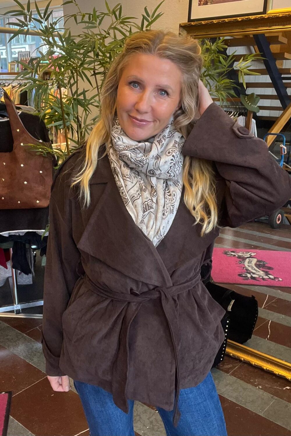 Kvinna i brun kappa och ljusmönstrad scarf poserar med ena handen i håret och ler mot kameran. Hon står i en klädbutik med klädställ, väska på stativ, stor växt och trappa i bakgrunden.