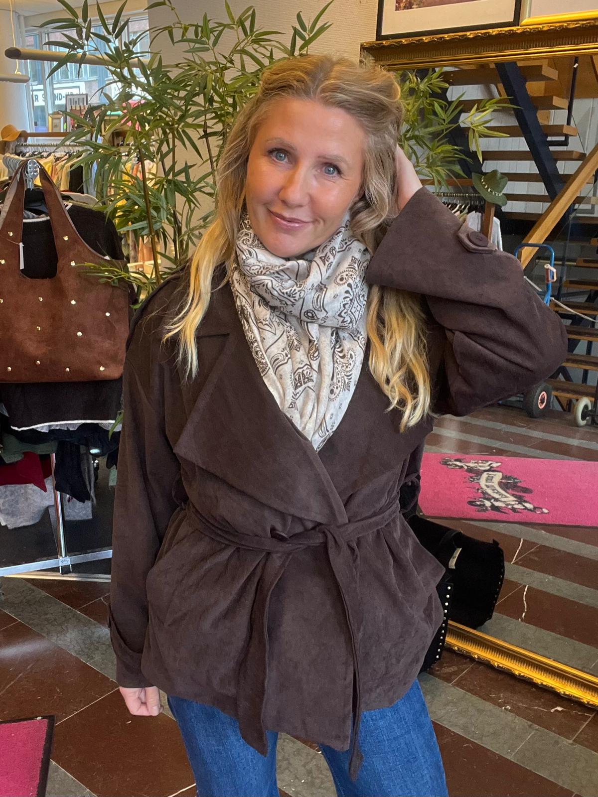 Kvinna i brun kappa och ljusmönstrad scarf poserar med ena handen i håret och ler mot kameran. Hon står i en klädbutik med klädställ, väska på stativ, stor växt och trappa i bakgrunden.
