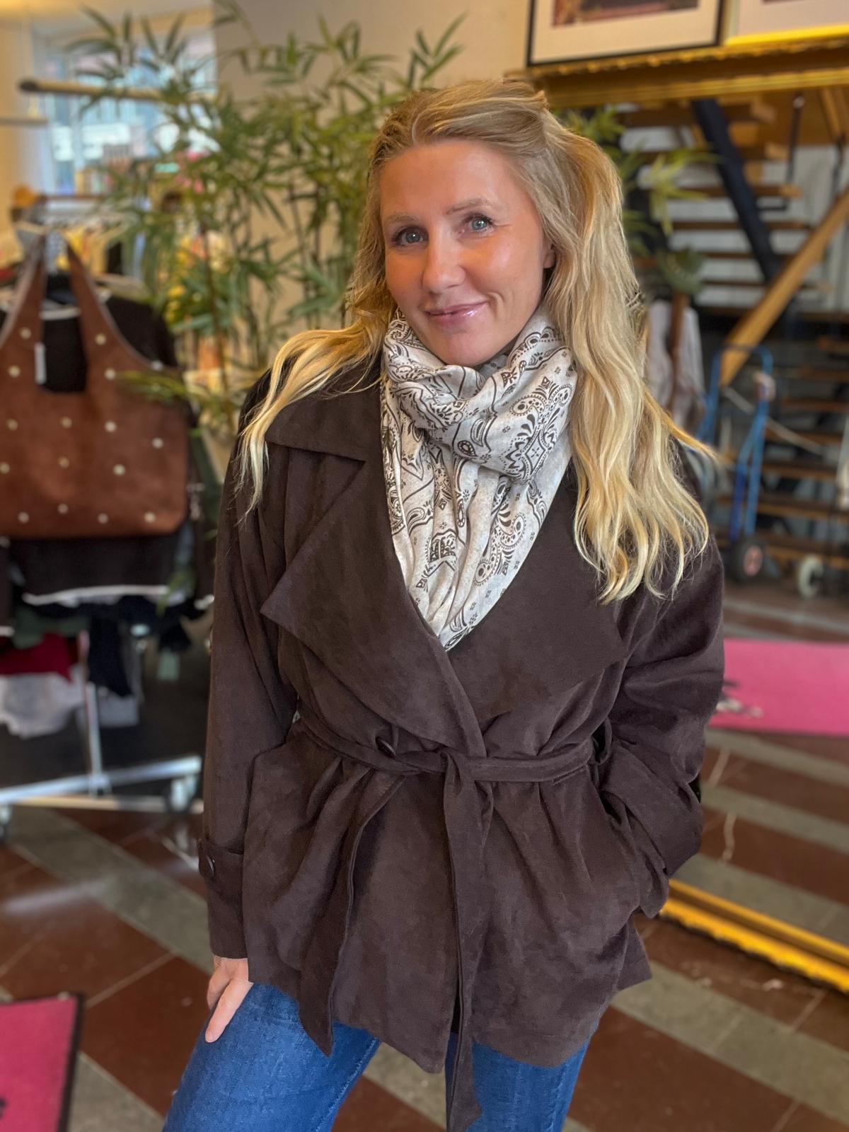 Leende kvinna med långt blont hår poserar inomhus, iförd brun knuten jacka och ljus mönstrad scarf. Hon står i en butikslik miljö med växter, en brun väska i bakgrunden och en trappa.