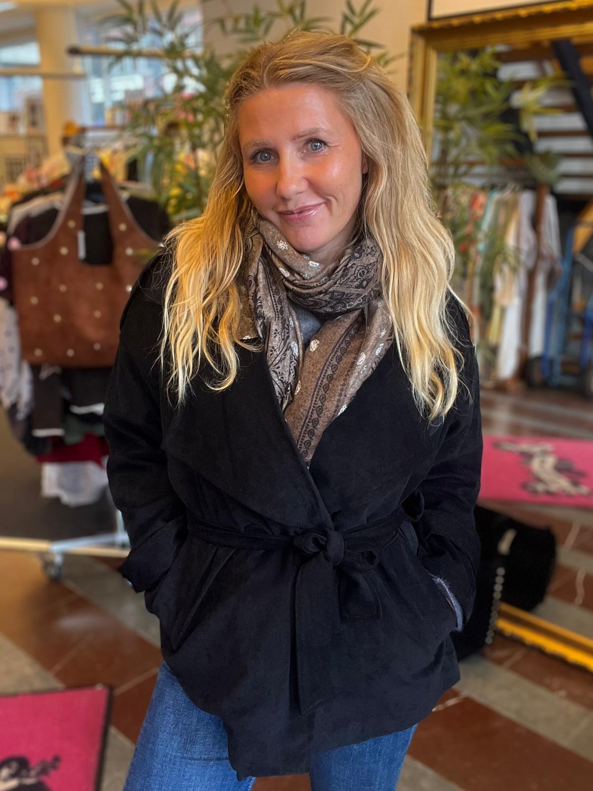 Blond kvinna står och ler mot kameran med händerna i fickorna, klädd i svart knuten jacka, mönstrad halsduk och jeans. Hon är inomhus i en butik med klädställningar och växter i bakgrunden.