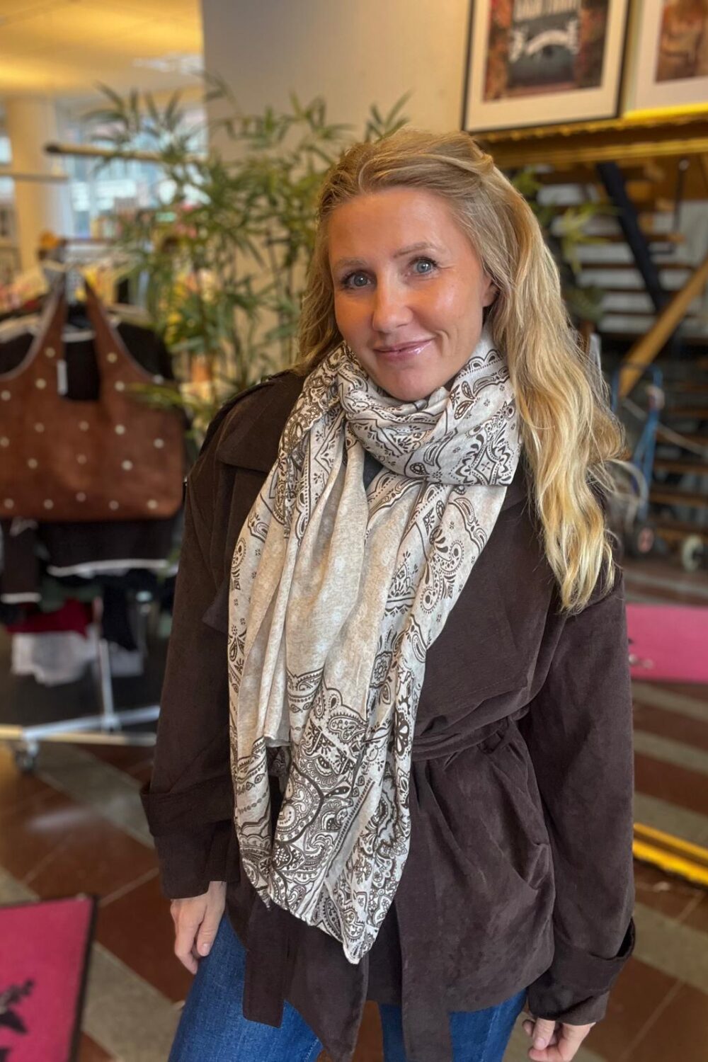 Blond kvinna står och ler mot kameran, klädd i brun jacka och ljus mönstrad sjal. Hon befinner sig i en butikslik miljö med växter och en klädställning med en brun väska i bakgrunden.