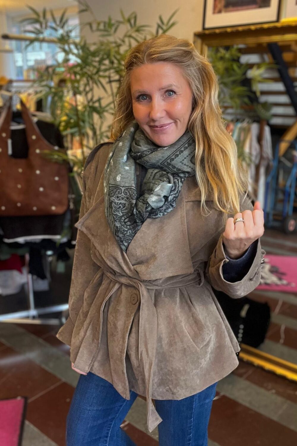 Leende kvinna poserar och håller upp handen med ring, klädd i brun knuten jacka, grå mönstrad scarf och jeans. Hon står inomhus i en butikslik miljö med växter, kläder och hyllor i bakgrunden.