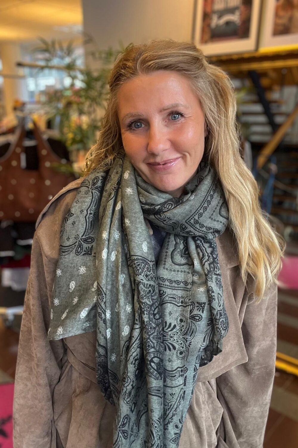 Leende kvinna med långt blont hår tittar in i kameran och står inomhus i en butik eller café. Hon bär en ljusbrun topp och en grå paisleymönstrad scarf; bakgrunden visar kläder och inredning.