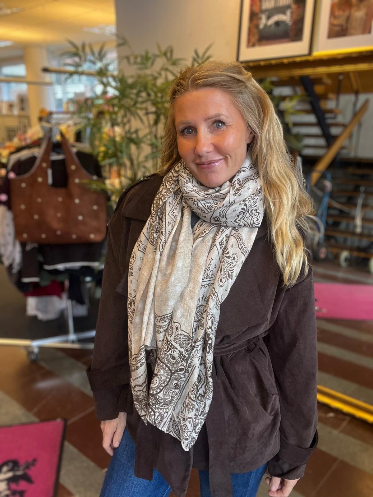 Blond kvinna står och ler mot kameran, klädd i brun jacka och ljus mönstrad sjal. Hon befinner sig i en butikslik miljö med växter och en klädställning med en brun väska i bakgrunden.