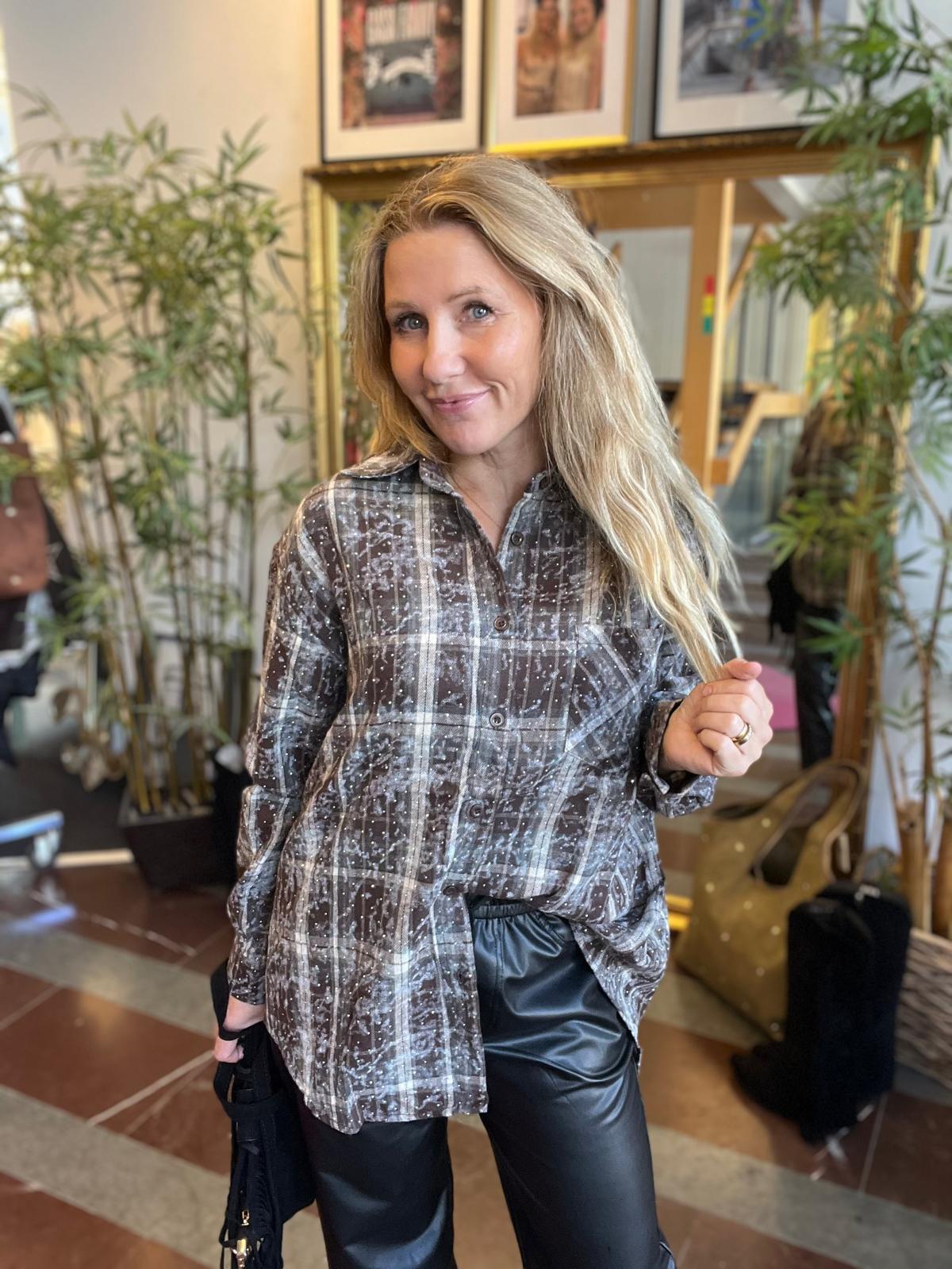 Blond kvinna står och ler mot kameran, håller en slinga av sitt hår och en svart väska. Hon bär en gråbrun rutig skjorta och svarta skinnbyxor, inomhus bland stora växter och inramade foton.