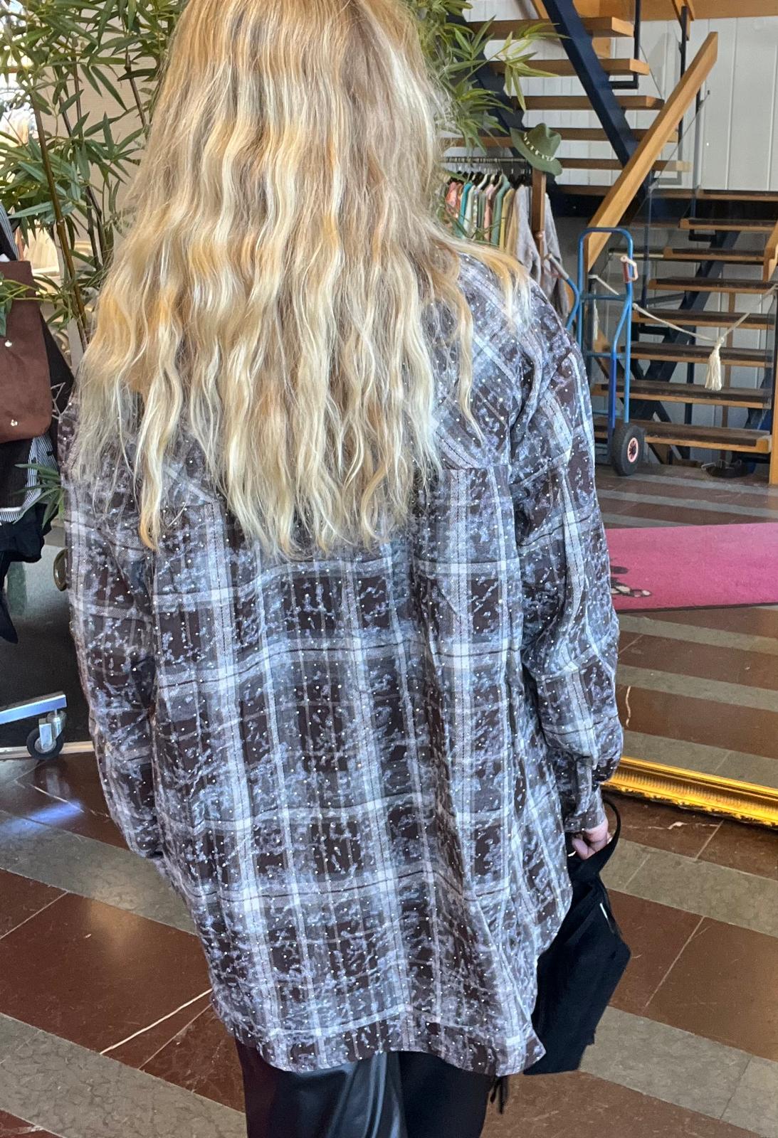 Person med långt, vågigt blont hår står med ryggen mot kameran och håller en svart väska. Personen bär en grå- och vitrutigt mönstrad skjorta. I bakgrunden syns klädställning, växt och trappa i en butiksmiljö.