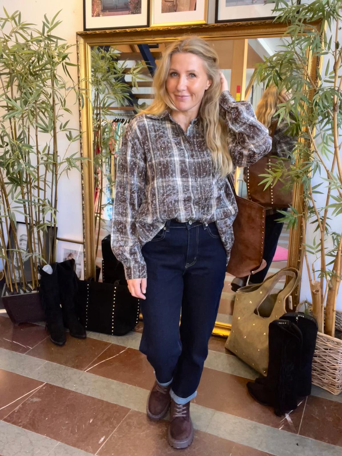 Blond kvinna poserar och ler framför en stor guldramsspegel, med ena handen i håret. Hon bär mönstrad blus, mörka jeans och bruna boots. Runt henne syns gröna växter, väskor och ett provrum/butiksmiljö.